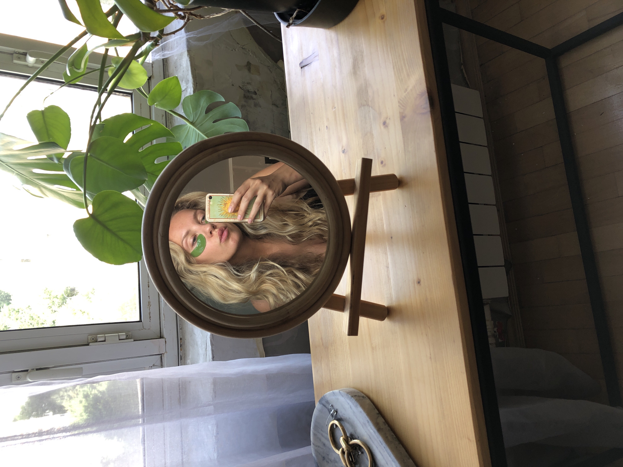 A young woman with blonde, wavy hair, wearing green under-eye masks, is taking a selfie in a round, wooden-framed mirror. She is holding a smartphone with a sun-themed tarot card case. The mirror reflects the woman's face, upper torso, and the phone. She is looking at the camera. The mirror is propped up on a wooden stand, placed on a light-colored wooden desk. To the left of the mirror is a marble tray with gold-colored jewelry, including earrings and a safety pin. Behind the mirror, a large Monstera plant is visible, with its leaves extending upwards and to the right. The scene appears to be indoors, near a window with white sheer curtains. The window frames the outside environment, which shows greenery and daylight. The desk has a dark metal frame underneath, and its surface shows wood grain and some faint marks. The reflection in the desk's surface is partially visible, showing floorboards and a light-colored object. The overall setting suggests a domestic environment, possibly a dressing table or a desk area. The specific location of Khimki, Russia, is not discernible from the visual elements of the image.