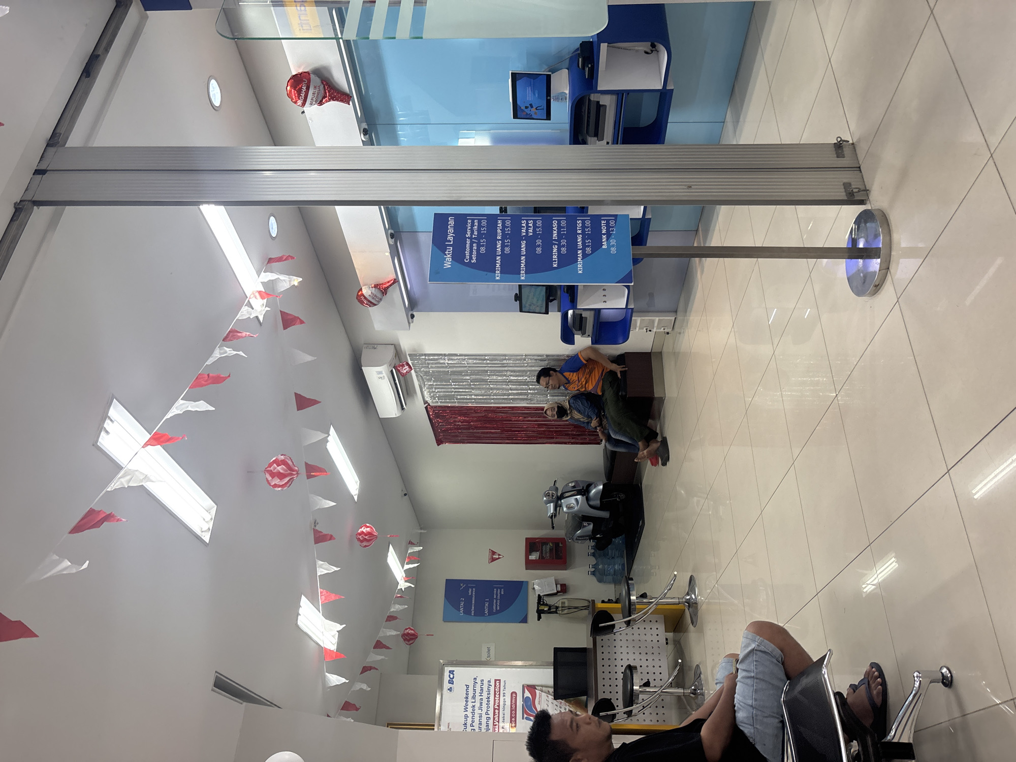 The image depicts the interior of a bank branch, likely in North Jakarta, Indonesia, as suggested by the "BCA" (Bank Central Asia) signage. The scene is decorated with red and white triangular flags strung across the ceiling and red and white spherical lanterns, indicating a festive or celebratory occasion, possibly related to Indonesian Independence Day. Several people are visible; a man in a black t-shirt and denim shorts is seated in the foreground on the left, facing away from the camera. In the middle ground, two individuals are seated on a bench to the right, one wearing a blue shirt and the other an orange shirt, both facing the counter. A motorcycle is parked behind them. The environment includes tiled floors, fluorescent lighting, and a service counter with computers and printers. A sign displaying "Waktu Layanan" (Service Hours) lists various banking services and their operating times. The overall atmosphere appears to be a typical bank waiting area, with some decorative elements adding a touch of local culture.