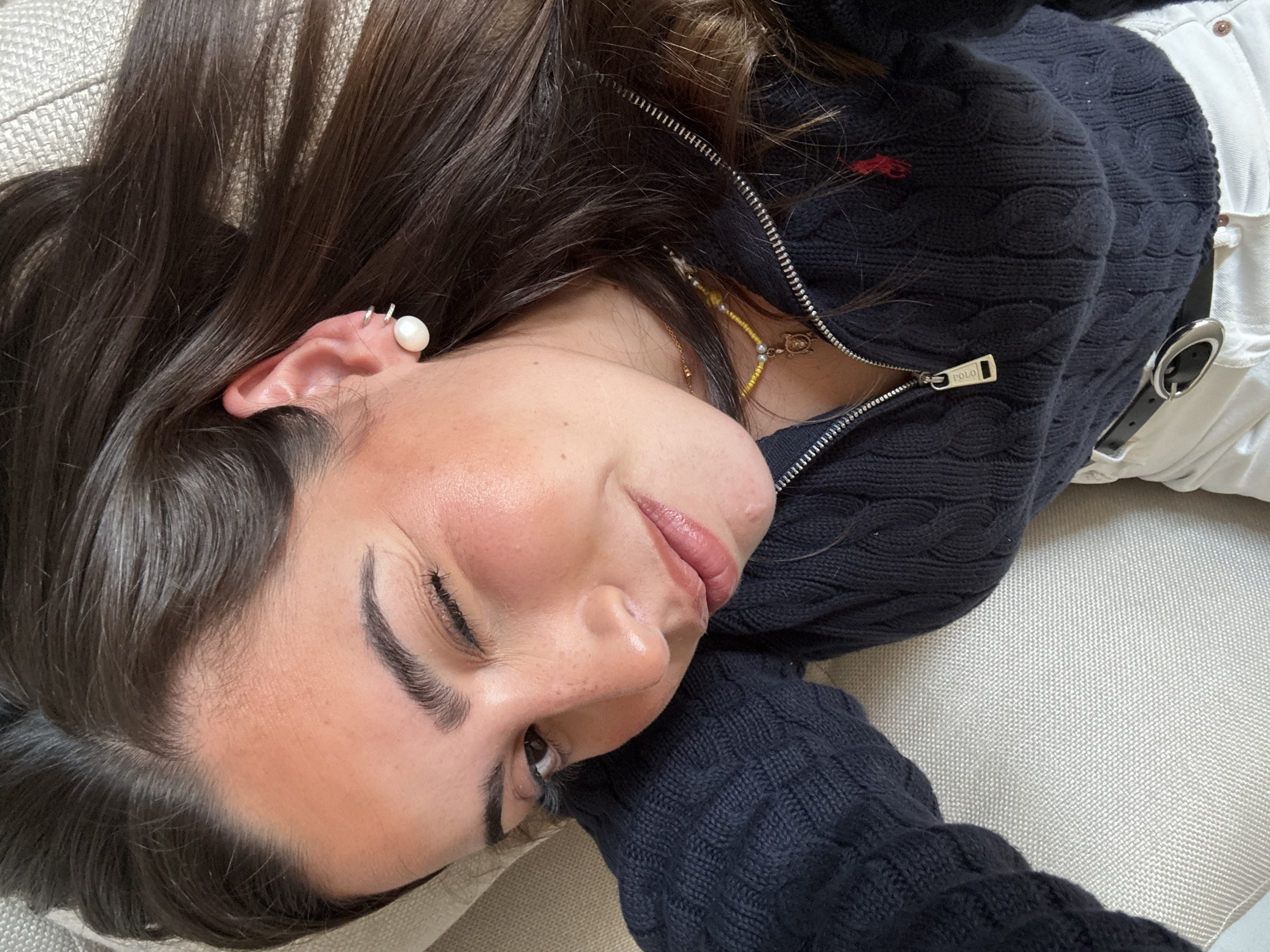 A young woman is lying on a light-colored upholstered surface. She is wearing a dark blue, cable-knit zip-up sweater with a red logo on the upper left chest and a Polo branded zipper pull. Underneath the sweater, a gold chain and a yellow and gold beaded necklace are visible. She also wears white pants with a black leather belt. She has dark brown hair and fair skin. A pearl earring is visible in her left ear, along with a smaller silver earring. The woman's eyes are mostly closed. The scene's location cannot be determined from the image.