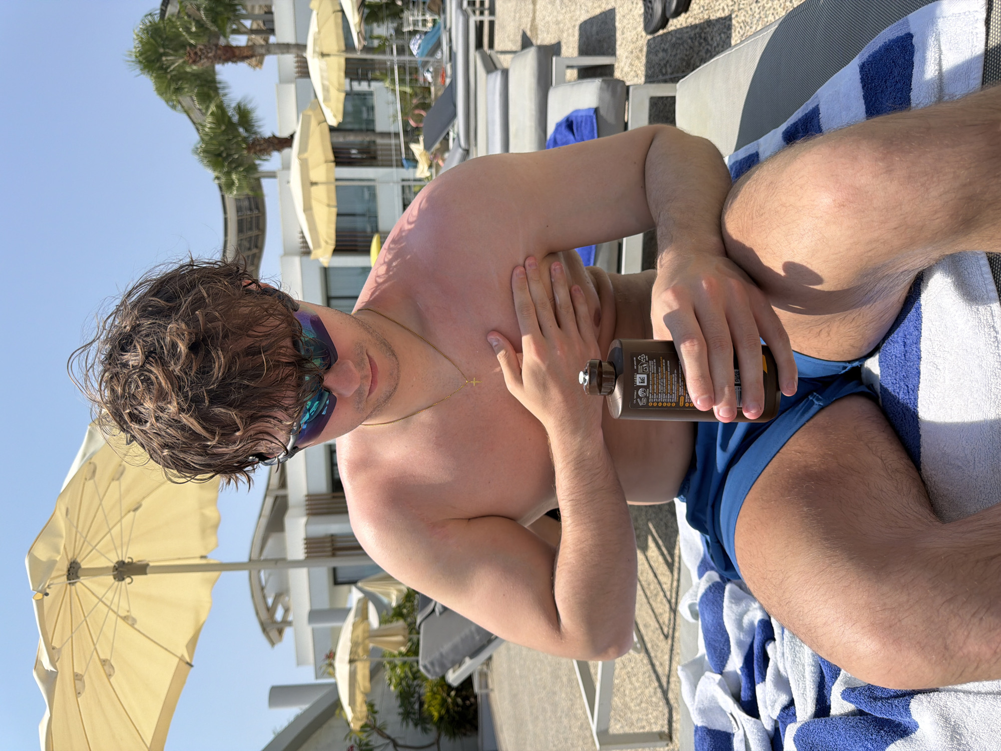 In Oroklini, Cyprus, a young man is sitting on a lounger by a pool, applying sunscreen to his shoulder. He has wavy brown hair, sunglasses with blue reflective lenses, and is wearing blue swim shorts. He is holding a brown bottle of sunscreen in his right hand and his left hand is on his shoulder. The background shows a bright blue sky, palm trees, and white buildings with umbrellas providing shade over lounge chairs. The overall scene is one of relaxation and protection from the sun.