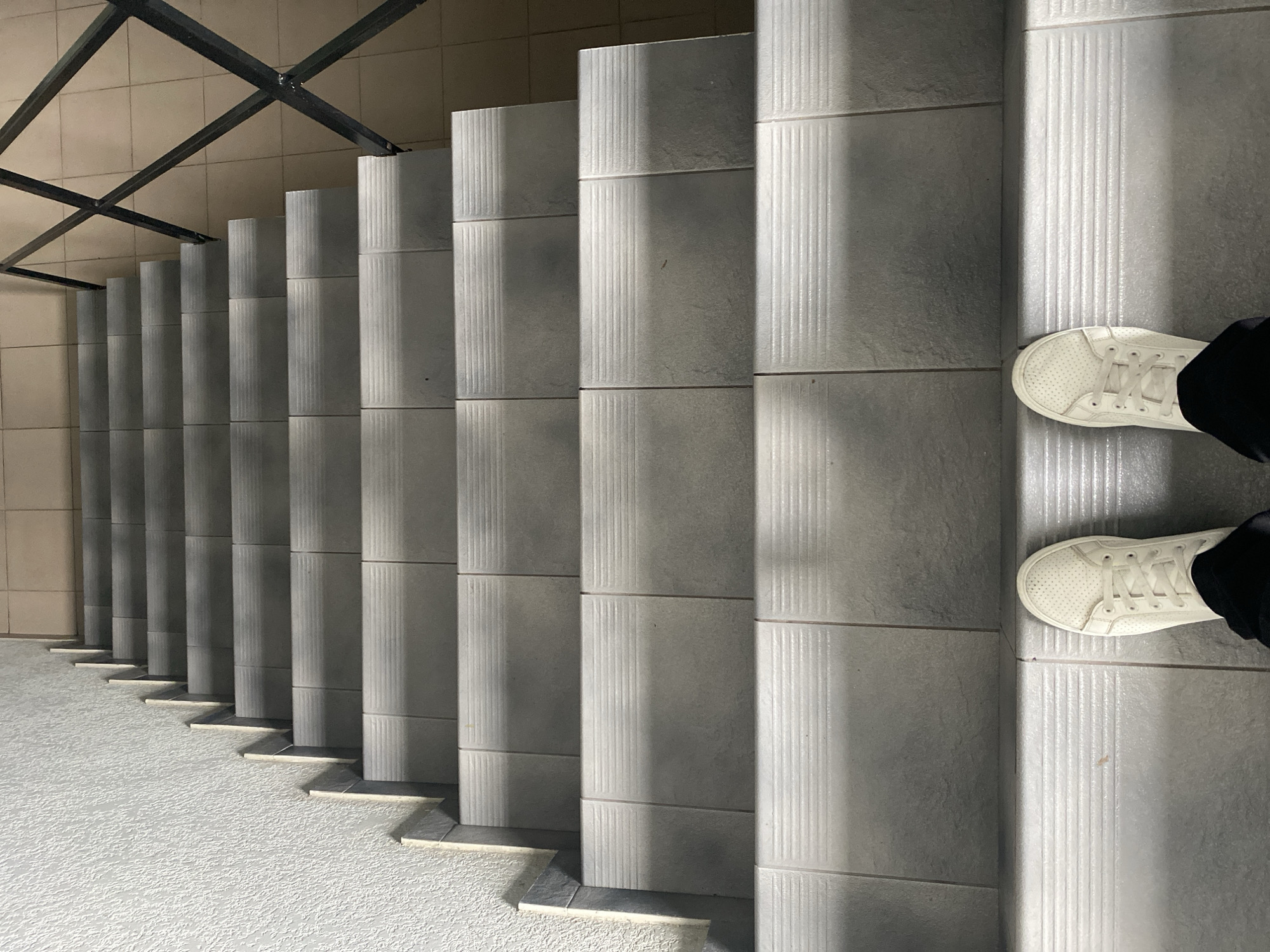The image shows a perspective view looking down a flight of grey tiled stairs. The stairs have a textured, horizontal pattern on their risers. On the left side of the stairs, there is a textured, light-colored wall. On the right side, a black metal railing with a geometric pattern is visible, and beyond that, the floor is tiled with beige squares. At the bottom of the stairs, a pair of white sneakers is visible, with black pants peeking out from beneath them. The lighting suggests an indoor environment, likely a stairwell. The location of Rozhny, Ukraine is not discernible from the visual information in the image.