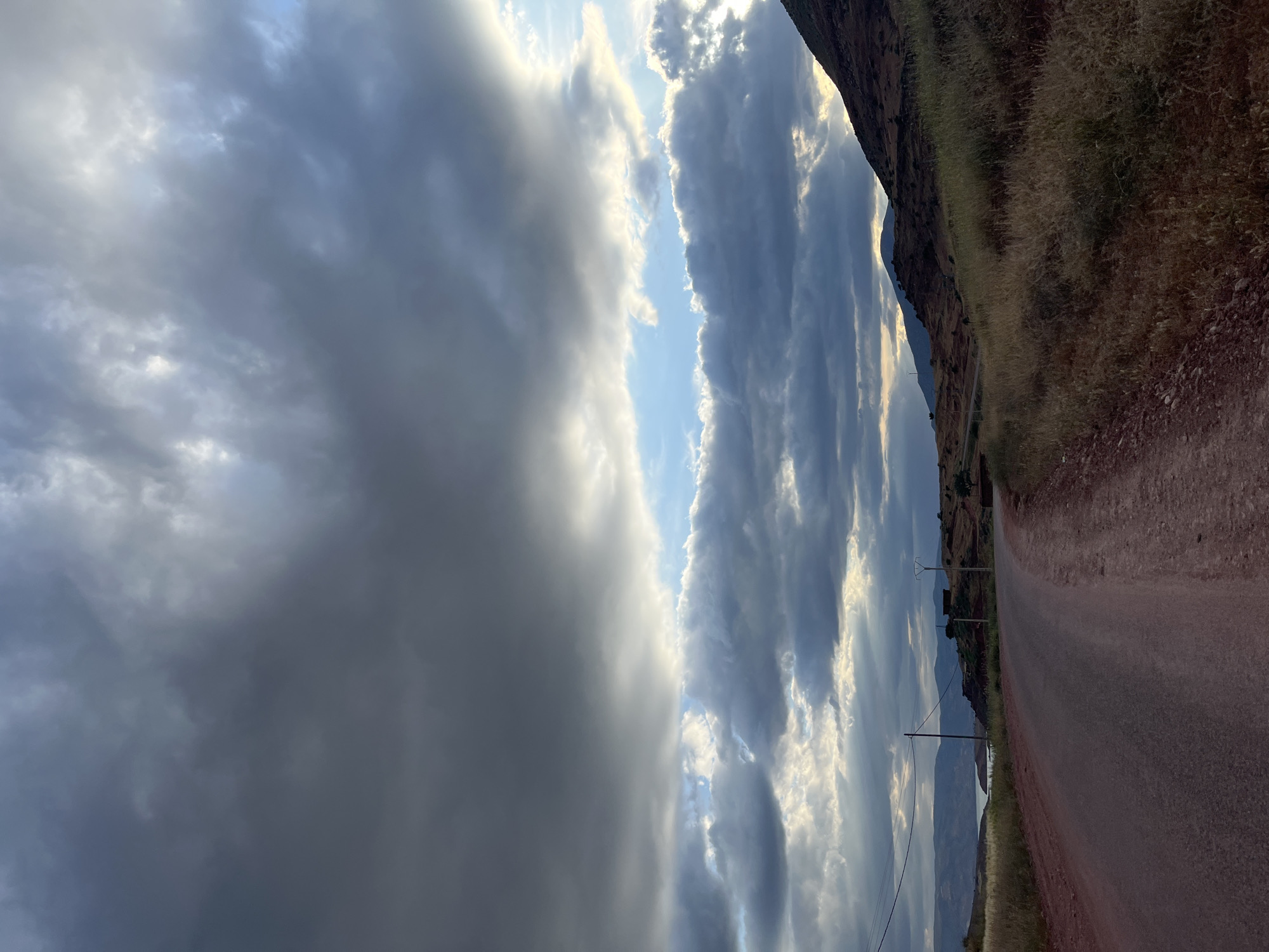 The image shows a dirt road in Ouaouizert, Morocco, with dramatic cloud formations in the sky. The road is red-brown and runs along the side of a rocky, grassy hill. Utility poles line the road, suggesting some infrastructure. In the background, there are hints of distant mountains. The sky is dominated by heavy, dark clouds on the left and a brighter, textured cloud formation with patches of blue sky in the middle. The overall scene suggests a rural or semi-rural area with striking natural features.