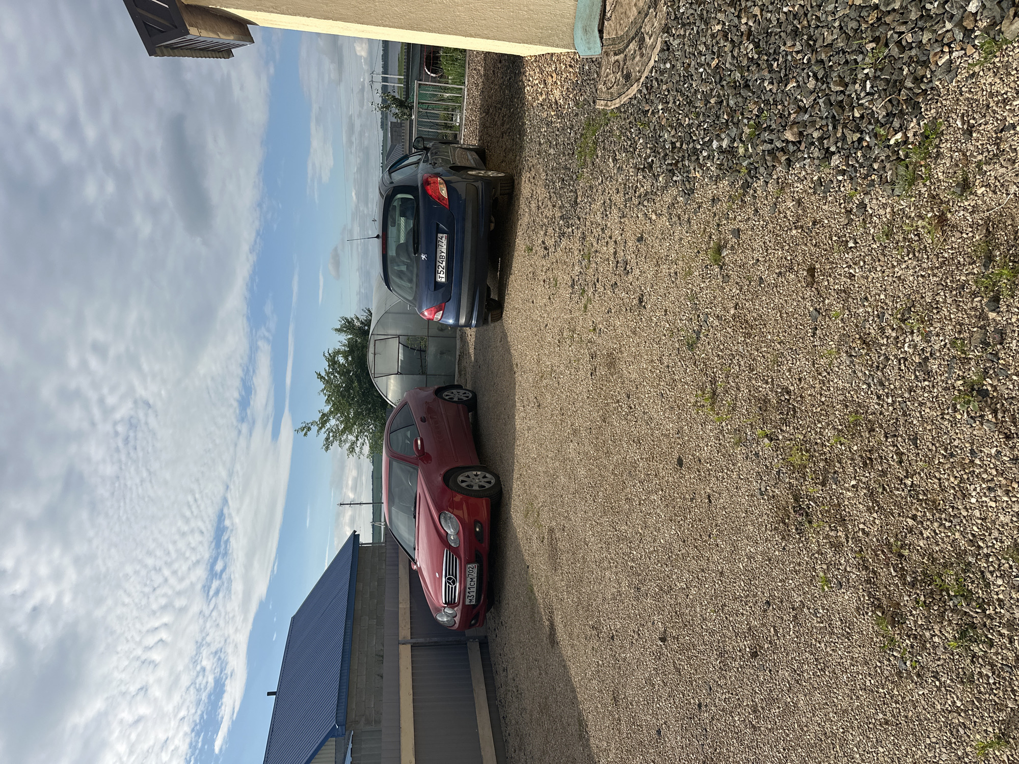 In Ozerne, Russia, a gravel lot is shown with two cars parked on it. The car on the left is a red Mercedes-Benz coupe, and the car on the right is a dark blue Peugeot hatchback. Behind the cars, a grey metal fence runs along the left side of the image. Beyond the fence, a building with a blue metal roof is visible, and further back, a greenhouse structure is present. The sky is partly cloudy with white cumulus clouds against a blue sky. To the right of the frame, a portion of a light-colored building is visible, and a patterned rug is partially visible on the ground.