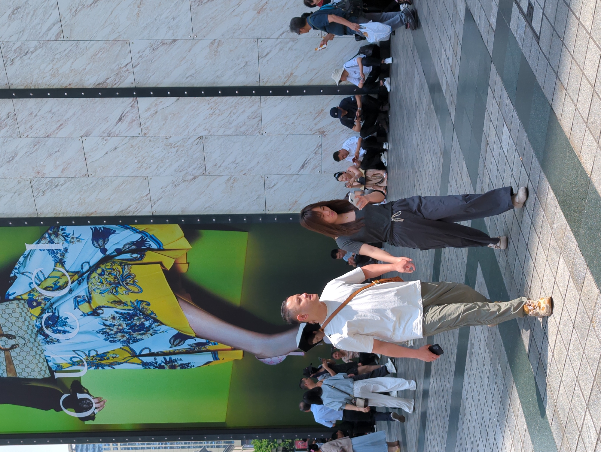 The image shows a scene in Yuzhong District, China. Several people are on a sidewalk next to a large advertisement for a fashion brand, likely Gucci. The people appear to be primarily of Asian descent, some sitting and some standing. Many seem to be looking at or taking pictures of the advertisement. A man is in the foreground, wearing a white shirt and khaki pants, seemingly posing in front of the ad. A woman in a gray outfit stands near him, possibly interacting with him. The advertisement features a large image of fashion items with yellow and blue patterns. The background includes a building with a marble-like facade and horizontal black metal strips. The sidewalk is made of gray tiles with a darker gray stripe pattern.