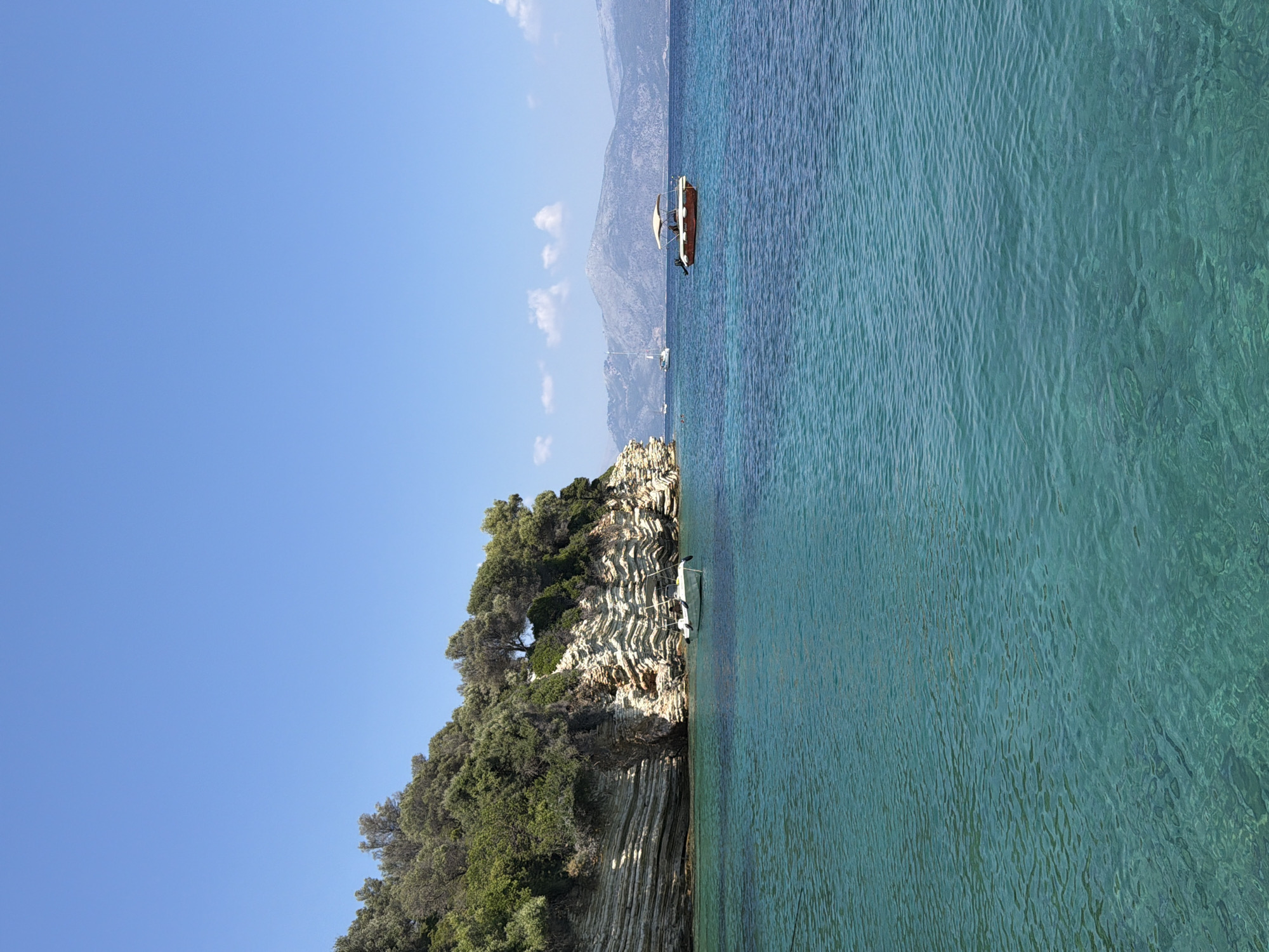 This is a vertical, eye-level, medium shot of a bright, clear day at sea near the Municipal Unit of Kastos, Greece. In the foreground, the crystal-clear turquoise water shimmers and ripples, with the seabed visible beneath the surface. To the left, a rocky cliff face covered in lush green trees rises steeply from the water. A small white boat is anchored near the base of the cliff. Further out in the sea, a small boat with a beige canopy and two people aboard is visible. In the background, distant mountains with a hazy, grayish hue are visible under a clear blue sky. A sailboat can be seen in the distance to the left of the boat with the canopy.