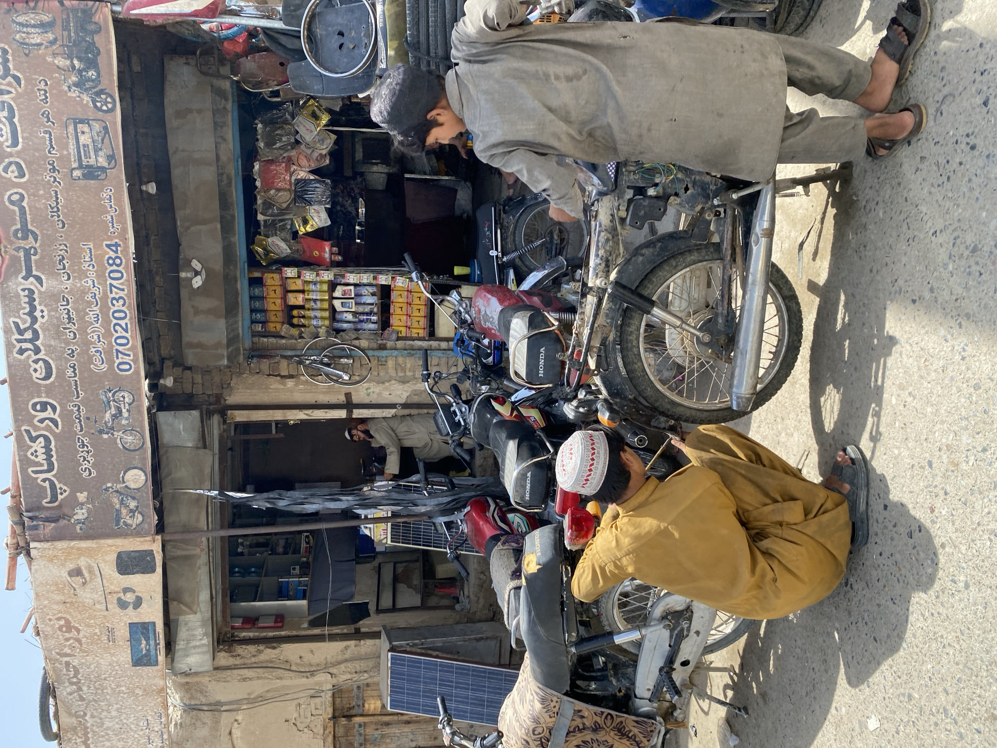 In Kandahar, Afghanistan, a street-level scene unfolds outside a motorcycle repair shop. The shop's facade features weathered signage with text in Pashto, and a faded depiction of motorcycles. Inside, shelves are stocked with various goods, including colorful bags and what appear to be engine oil cans. Multiple motorcycles, some in various states of repair, are parked on the street.
Two individuals are actively engaged with the motorcycles. One, wearing a traditional mustard-yellow shalwar kameez and a patterned cap, is crouched down, appearing to work on the engine of a motorcycle. The other, a younger person in a grey shalwar kameez, stands and is also focused on a motorcycle. A third individual, a man in a light brown shalwar kameez and a white cap, is visible inside the shop, looking towards the right of the frame.
The environment is dusty, with a sun-drenched street and a clear sky. Solar panels are visible against one of the shop's walls, and the overall impression is one of a busy, working-class commercial area in the city.