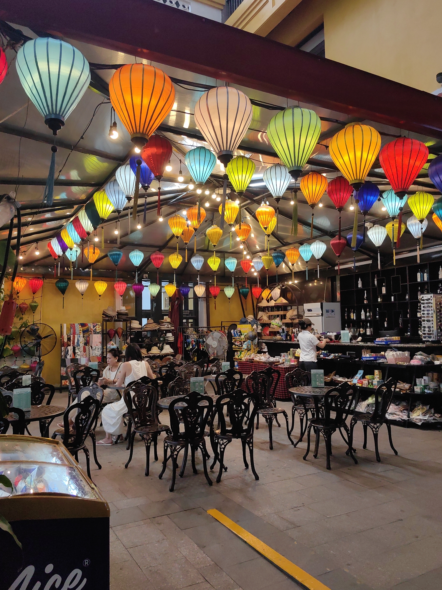 An indoor setting, possibly a restaurant or shop, in Bai Chay Ward, Vietnam is filled with colorful lanterns hanging from the ceiling. Tables and chairs occupy the foreground. Two women are seated at a table. A person wearing a white shirt is standing behind a counter. Shelves containing various products line the background.