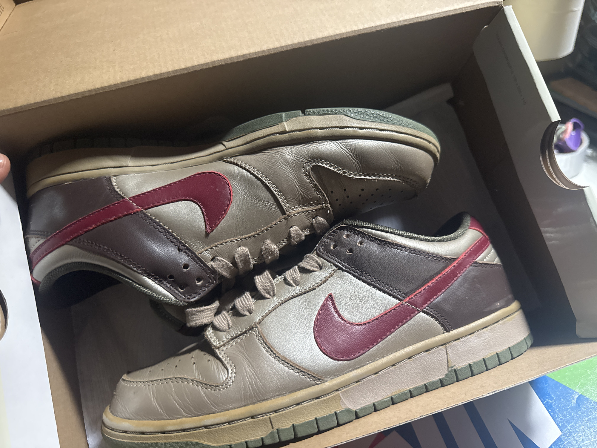 The image shows a pair of used Nike Dunk Low sneakers in a cardboard shoebox. The sneakers have a brown and tan color scheme with red Nike swooshes. The soles and midsoles are greenish, suggesting wear and age. The laces are also a light tan color. The shoebox is plain brown cardboard. No specific location in Barcelona, Spain, is discernible from the image.