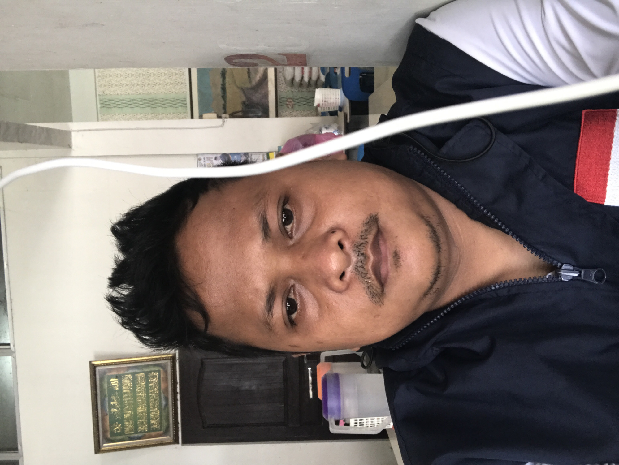 A person is visible wearing a dark blue jacket with a white and red section on the right shoulder, presumably indoors based on the background. The background includes a light-colored wall with framed artwork and various items on a shelf. The image seems to be taken at an angle and partially obscured by a white wire. There is no specific information to confirm that the location is in the city of Grong-Grong, Indonesia.