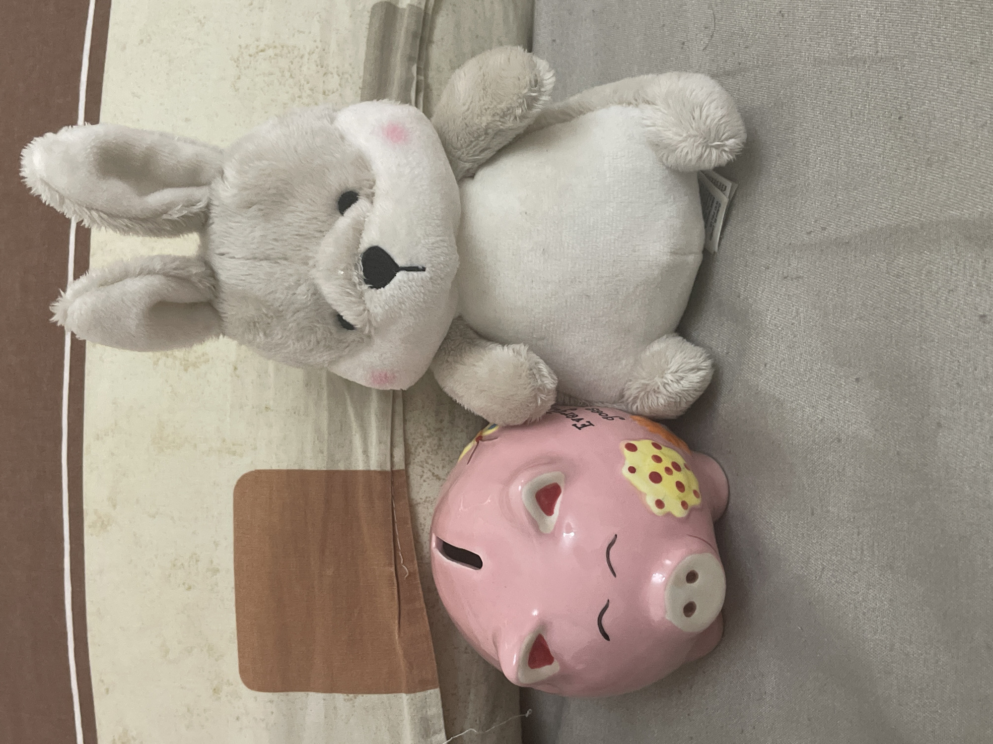 This scene, located in the city of Ihtiman, Bulgaria, depicts two static objects positioned on a light grey, textured fabric surface. On the left, a pink ceramic piggy bank is oriented with its front facing forward. It features a horizontal coin slot on its top surface, two painted, closed-eye arches, a circular snout with two nostrils, and two pointed ears with red inner sections. A yellow, irregular shape with red circular spots is painted on its left side. Partial black text, "Ever" and "goes," is visible on its body. To the right of the piggy bank, a light grey plush toy, consistent with a rabbit, is in a seated position. This toy has two prominent, upright ears, small black eyes, a black heart-shaped nose, and two small pink blush marks on its cheeks. A white manufacturer's tag is partially visible beneath the toy's left foot. The background consists of a light beige-cream fabric with a subtle mottled pattern. A distinct brown rectangular patch with rounded corners is present on the upper left of the background, with a horizontal band of a thin white line and a wider brown line positioned above it.