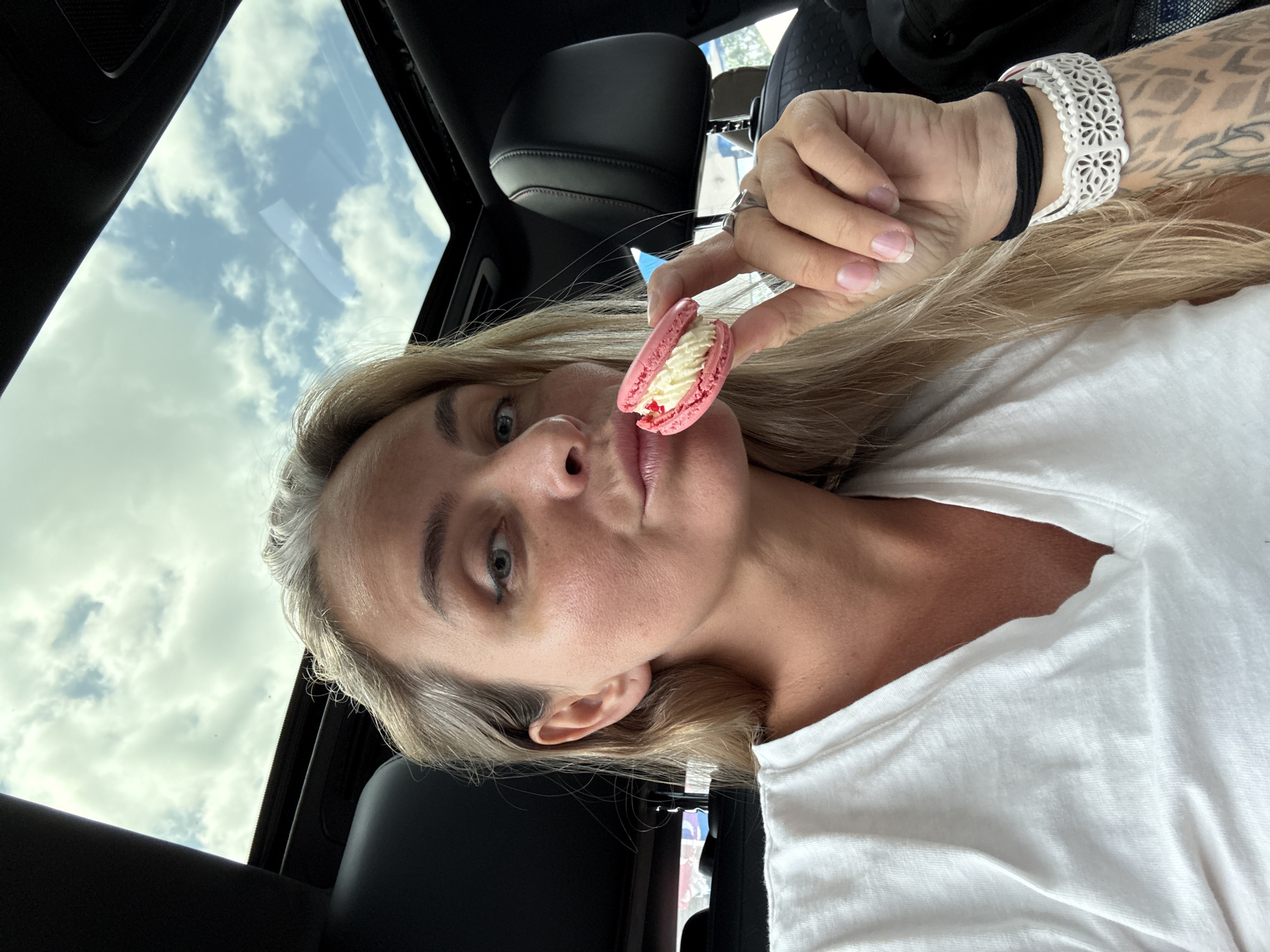 A woman is sitting in a car with a sunroof open, looking at the camera and holding a pink macaron with white cream filling. She is wearing a white V-neck t-shirt and has blonde hair. Her left hand has a ring and a white and black bracelet. She has tattoos on her left forearm. The interior of the car is visible, with black leather seats and the ceiling. The sky with clouds is visible through the sunroof. The image does not provide any specific information about the location in the city of Tha Uthae, Thailand.