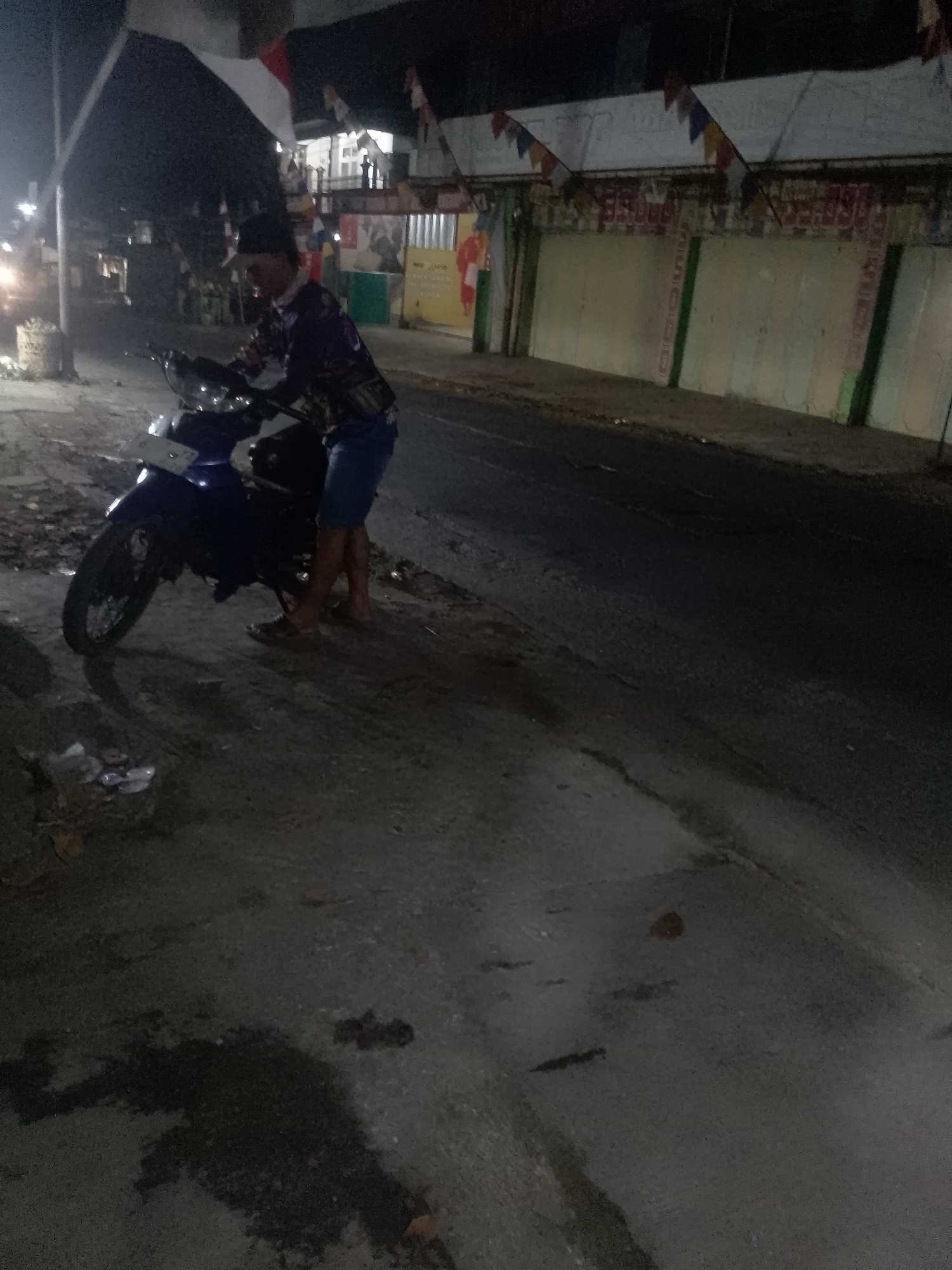 In the city of Cikalongkulon, Indonesia, at night, a person is standing next to a blue motorcycle. The person is wearing a dark colored long-sleeved shirt with a pattern and blue shorts. They are also wearing a dark hat and sandals. The motorcycle is parked on a paved surface, likely a street or roadside, with debris and puddles visible. In the background, there are buildings with awnings decorated with small flags. The lighting suggests it is an urban or semi-urban area at night.