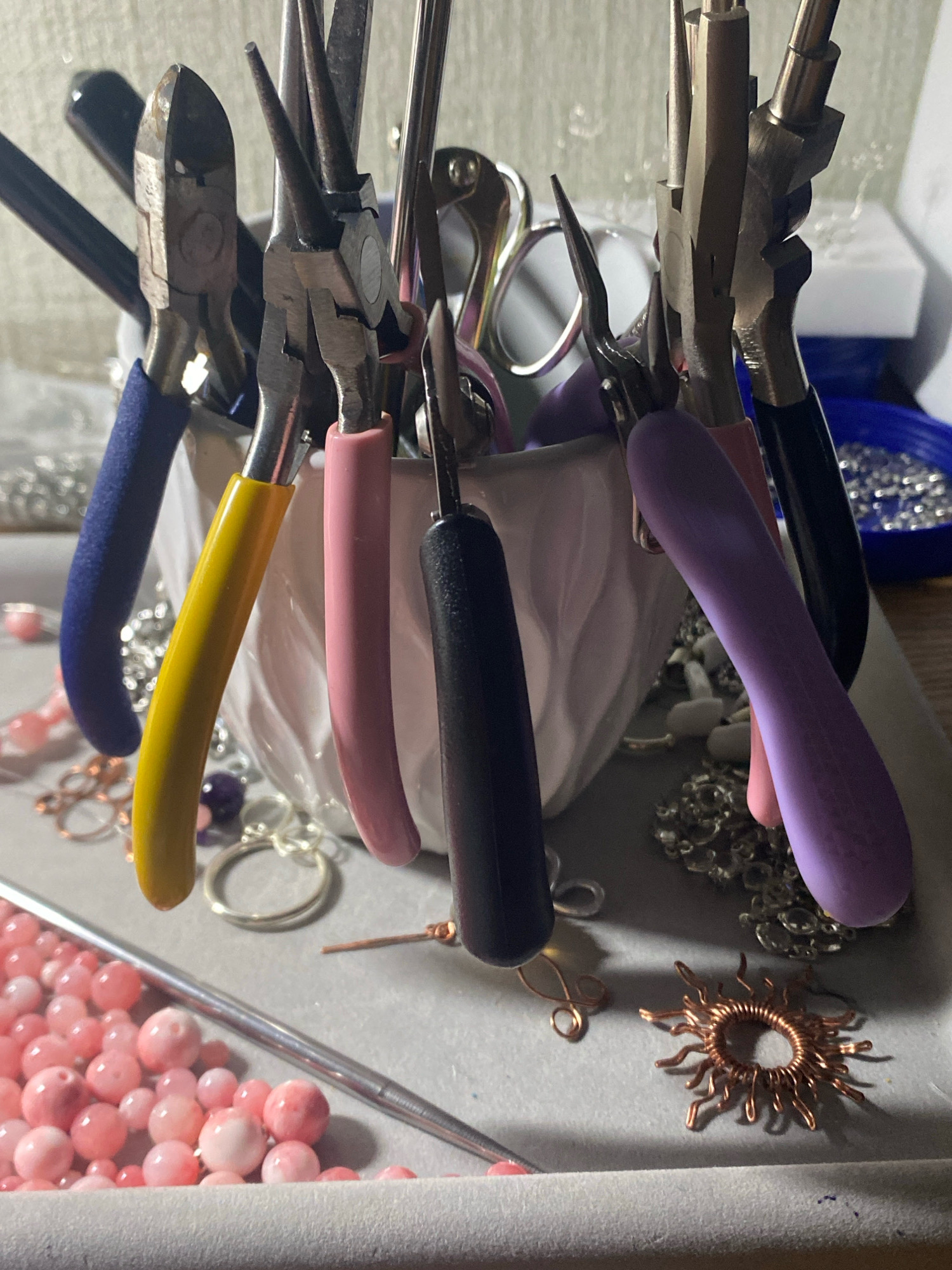 The image shows a collection of jewelry-making tools and supplies, including various pliers with colored handles, beads, and wire findings. The tools are arranged in a white ceramic container, and some appear to be used for cutting, bending, and shaping wire. Beads in shades of pink and purple are scattered around the tools, along with metal rings and a wire sun charm. The background is blurred, but suggests a crafting or workspace environment. The image is well-lit, highlighting the details of the objects. No specific location in Kushchovskaya, Russia, can be identified from the image.
