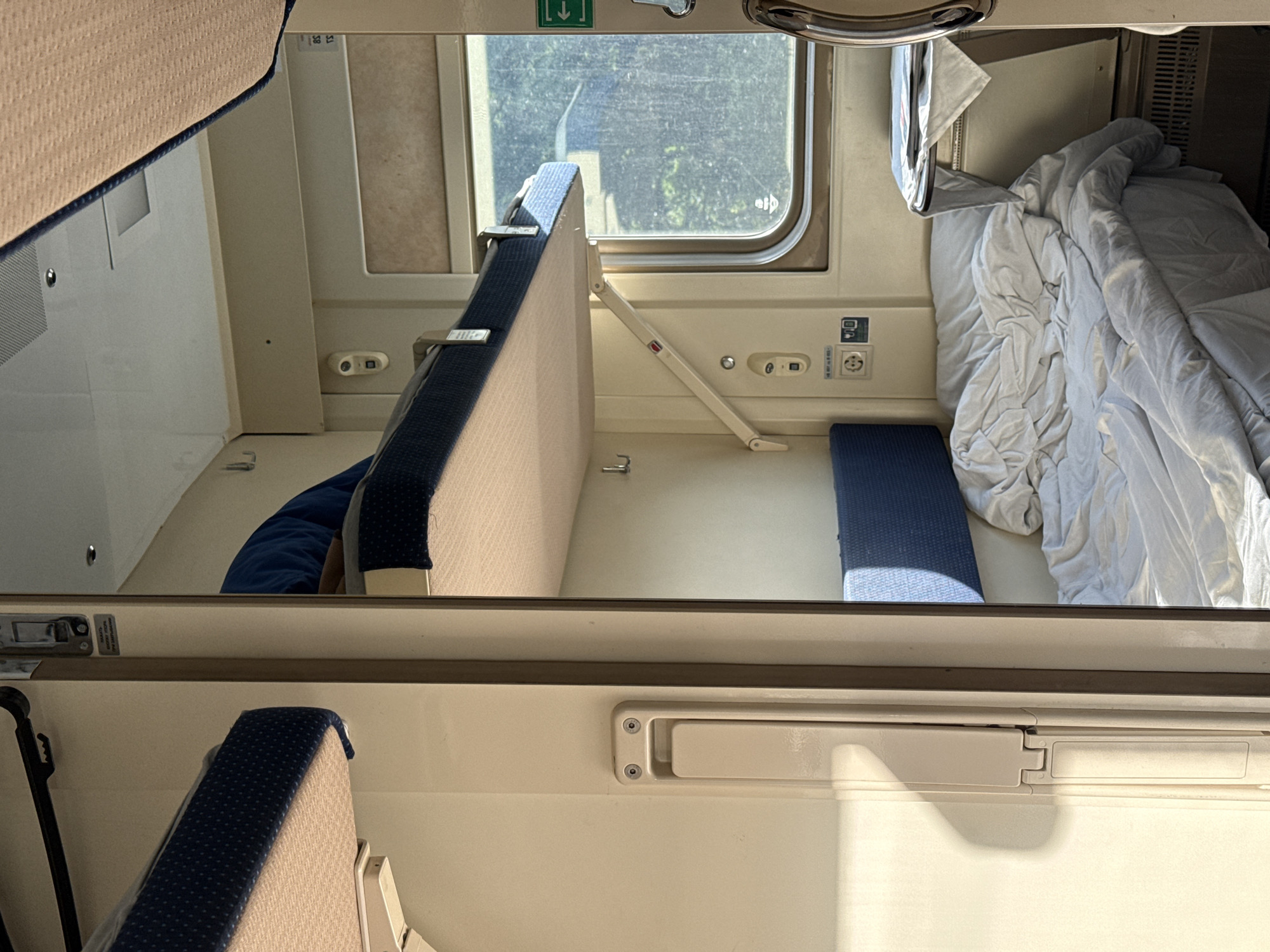 The image depicts the interior of a sleeper cabin on a train. The scene is brightly lit by daylight filtering through a window. The cabin contains two berths: an upper berth partially visible at the top of the frame and a lower berth that is configured as a seating area. A blue cushion rests on a small section of the floor beside the lower berth. A white comforter is rumpled on the right side of the lower berth. The opposite side of the lower berth has a hinged seat back in the upright position. The cabin wall features electrical outlets, a window with a partially obscured view to the outside, and a ventilation vent. The location in the city of Kirpichnoye, Russia, is not evident from the image.