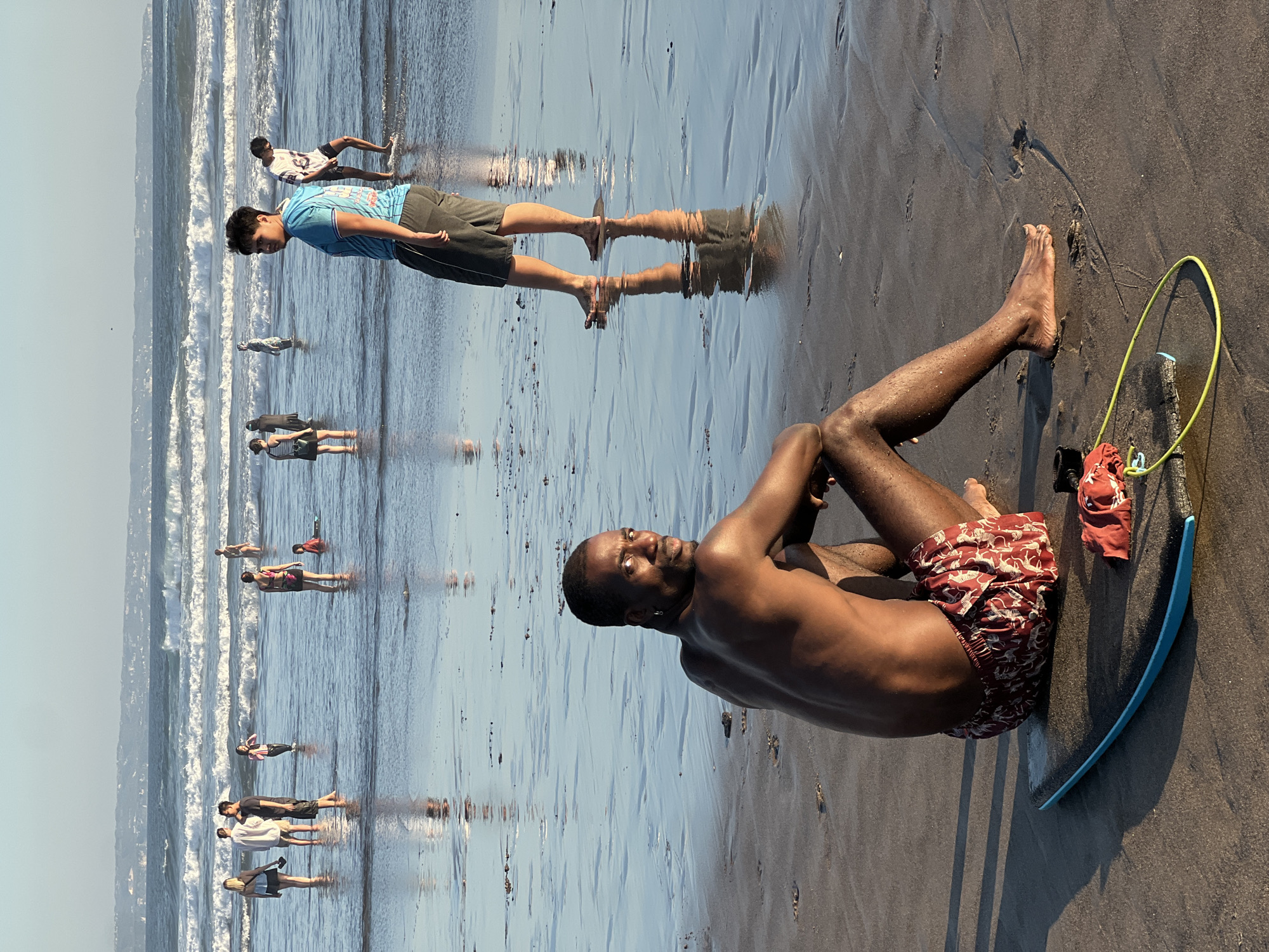 In the city of North Kuta, Indonesia, this image captures a serene beach scene with people enjoying the ocean and sand. In the foreground, a man sits on the wet sand, looking back at the camera with a slight smile. He is shirtless, wearing patterned red shorts, and is positioned next to a boogie board. Further out, several individuals are visible in the shallow water, some wading and others standing. The ocean exhibits gentle waves, and the sky is a clear, pale blue. The wet sand reflects the sky and the figures, creating a sense of depth and tranquility. The overall atmosphere suggests a peaceful day at the beach.