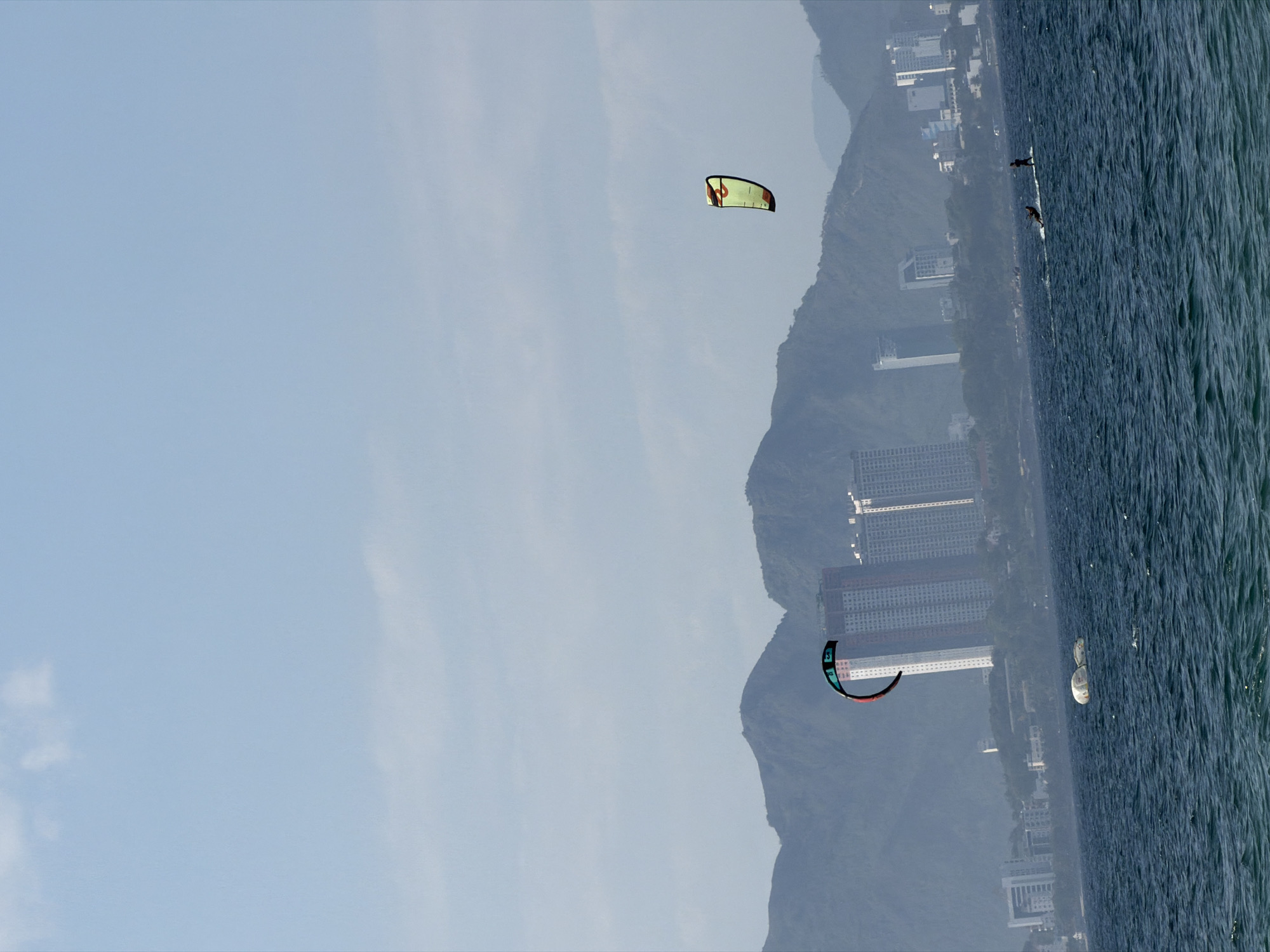 The image captures a coastal scene in Nha Trang Ward, Vietnam, under a hazy, light blue sky with faint cloud formations. The foreground consists of dark blue-green ocean water, agitated with visible surface choppiness and waves.
Two individuals are engaged in watersports on the ocean. On the right, a person stands on a light-colored board, possibly a paddleboard, moving across the water. To the left, another person or object is partially submerged or on a board, associated with a large, predominantly black kite with red, green, and blue accents, positioned high in the mid-left portion of the sky. A second, smaller kite, colored yellow and green, is visible further right and higher in the sky. Two distinct white, rounded objects, resembling buoys or inflatable markers, float on the water in the mid-left.
The midground features the urban landscape of Nha Trang. Numerous multi-story buildings, including prominent tall skyscrapers with rectangular silhouettes and visible window grids, line the coast. These structures display colors ranging from white and light grey to beige. A range of mountains, appearing in subdued grey-green hues due to atmospheric haze, forms the distant background, rising behind the city's buildings. The scene is illuminated by natural daylight.