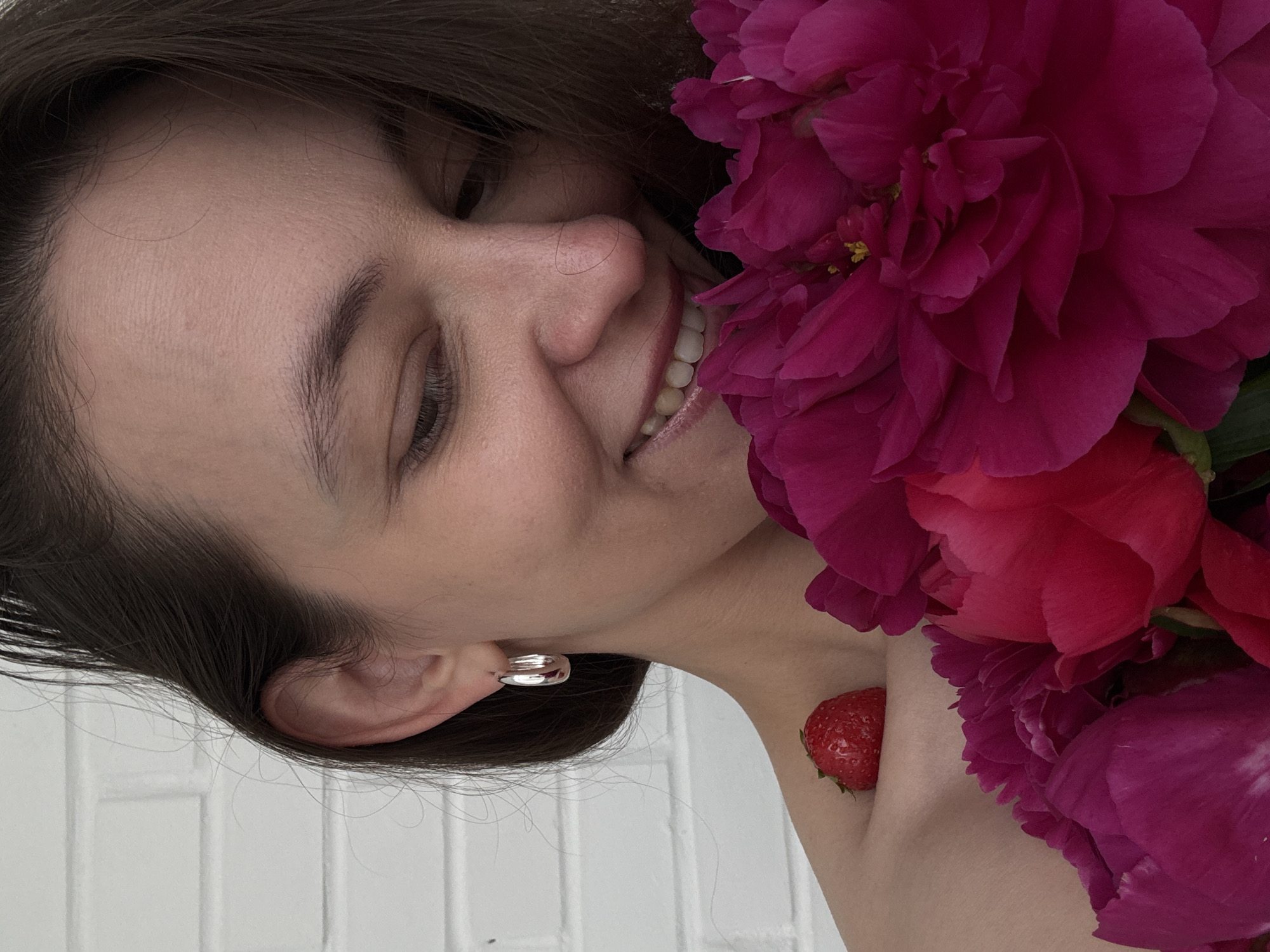 The scene is located in Chaikovsky, Russia. It features a close-up portrait of an adult female with dark brown hair, positioned against a white brick wall background. The woman is smiling broadly with a closed-lip expression, her head tilted downwards and to her right, and her eyes looking in the same direction. Her left ear is visible, adorned with a silver-colored, reflective, chunky hoop earring. Her forehead exhibits fine lines, and stray wisps of dark hair are visible around her face and ear. A single ripe red strawberry with visible seeds rests on her bare left shoulder/clavicle area. A large bouquet of dark pink/magenta peonies, characterized by layered petals, occupies the lower right and central portion of the frame, partially obscuring her lower face and chest. Some yellow stamens are visible in the center of one of the peony blooms. The lighting is soft and even, highlighting skin texture and flower details.