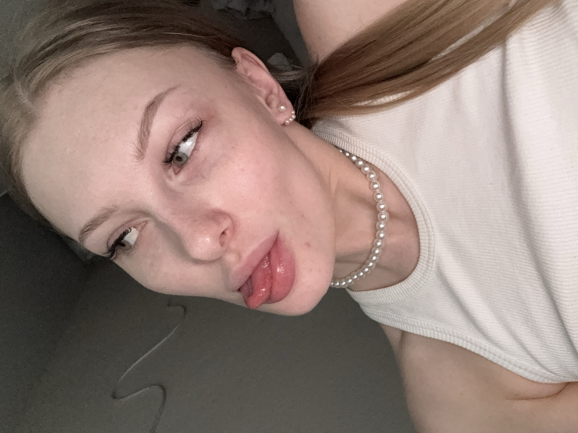 A fair-skinned woman is seen in a close-up, angled shot. She has blonde hair pulled back in a low ponytail. Her features include light eyebrows, visible eyelashes, and light-colored eyes. Her tongue is sticking out. She is wearing a white ribbed tank top and a pearl necklace. She is wearing pearl earrings. The background is out of focus. There is no evidence to indicate this photograph was taken in Perm, Russia.