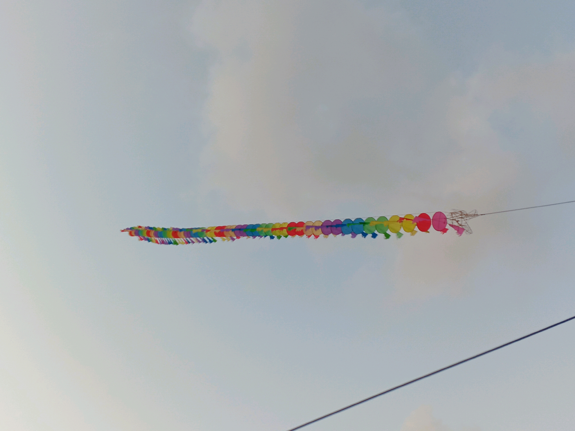A long, colorful string of circular ornaments, resembling a garland or a kite tail, is suspended horizontally against a pale, overcast sky. The ornaments are connected in a chain, displaying a vibrant spectrum of colors including red, orange, yellow, green, blue, and purple, arranged in a repeating pattern. At the far right end of the garland, there appears to be a small, more complex kite-like structure, possibly made of lighter material, with some delicate lattice work visible. A thin, dark wire runs diagonally across the lower right portion of the image, suggesting the presence of electrical or communication lines. The overall scene conveys a festive or decorative element set against a neutral, atmospheric backdrop.