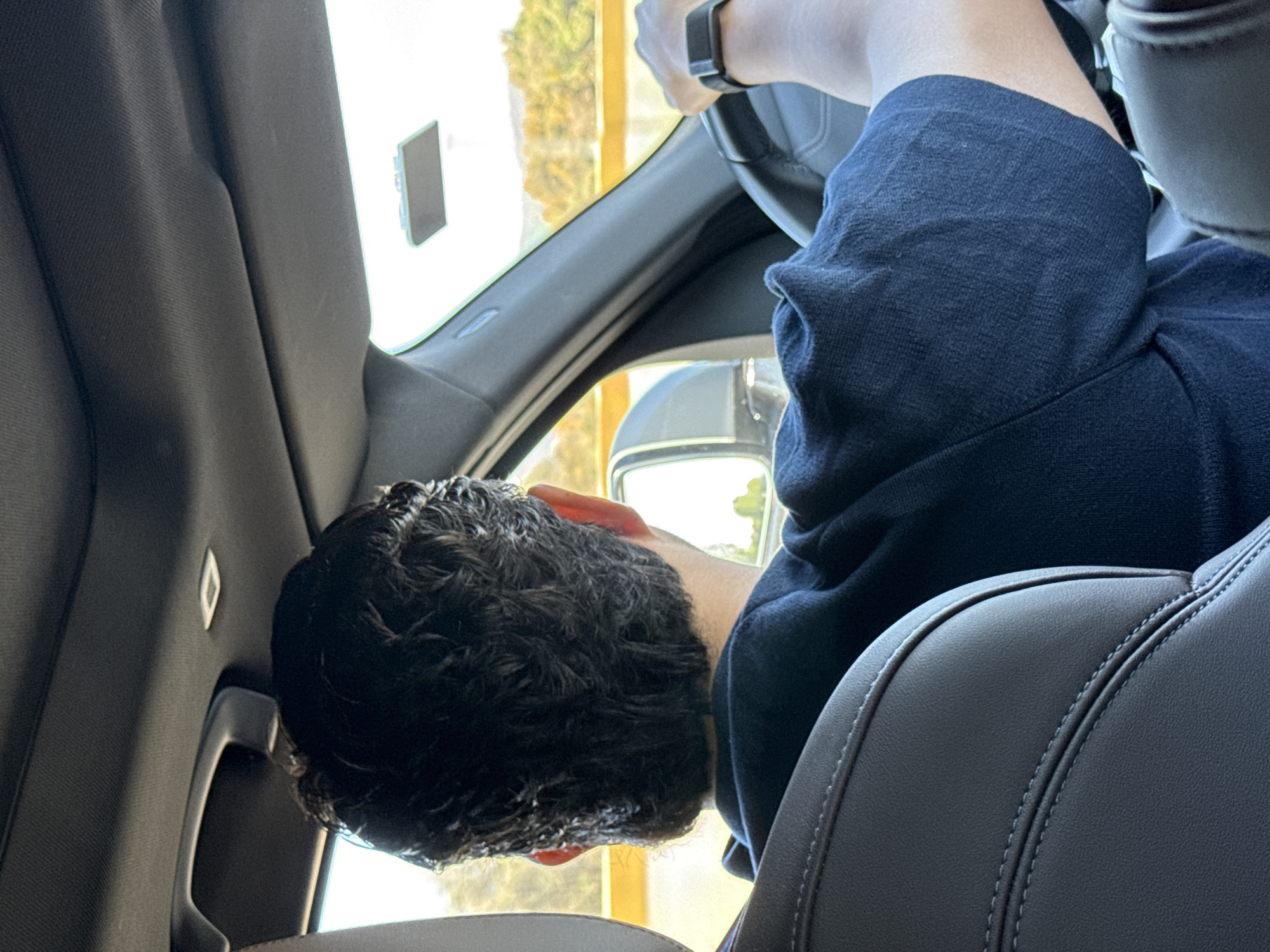 The image displays the interior of a vehicle, viewed from a rear passenger seat, looking forward at the driver from behind. The driver is a male with dark, curly hair, wearing a dark blue, short-sleeved shirt. His left arm is visible, with a black-banded smartwatch on his wrist, as his hands grip a dark-colored steering wheel. The vehicle's interior features a dark headliner with a small rectangular overhead light fixture. The back of the driver's seat, visible in the foreground, is dark gray/black leather with visible stitching. Through the front windshield and the driver's side mirror, a bright, sunny daytime outdoor environment is visible, characterized by a blurry yellow road barrier and distant landscape with yellowish-brown vegetation or hills under a bright sky, indicative of a highway or open road. The scene is situated in San Matías Tlalancaleca, Mexico.