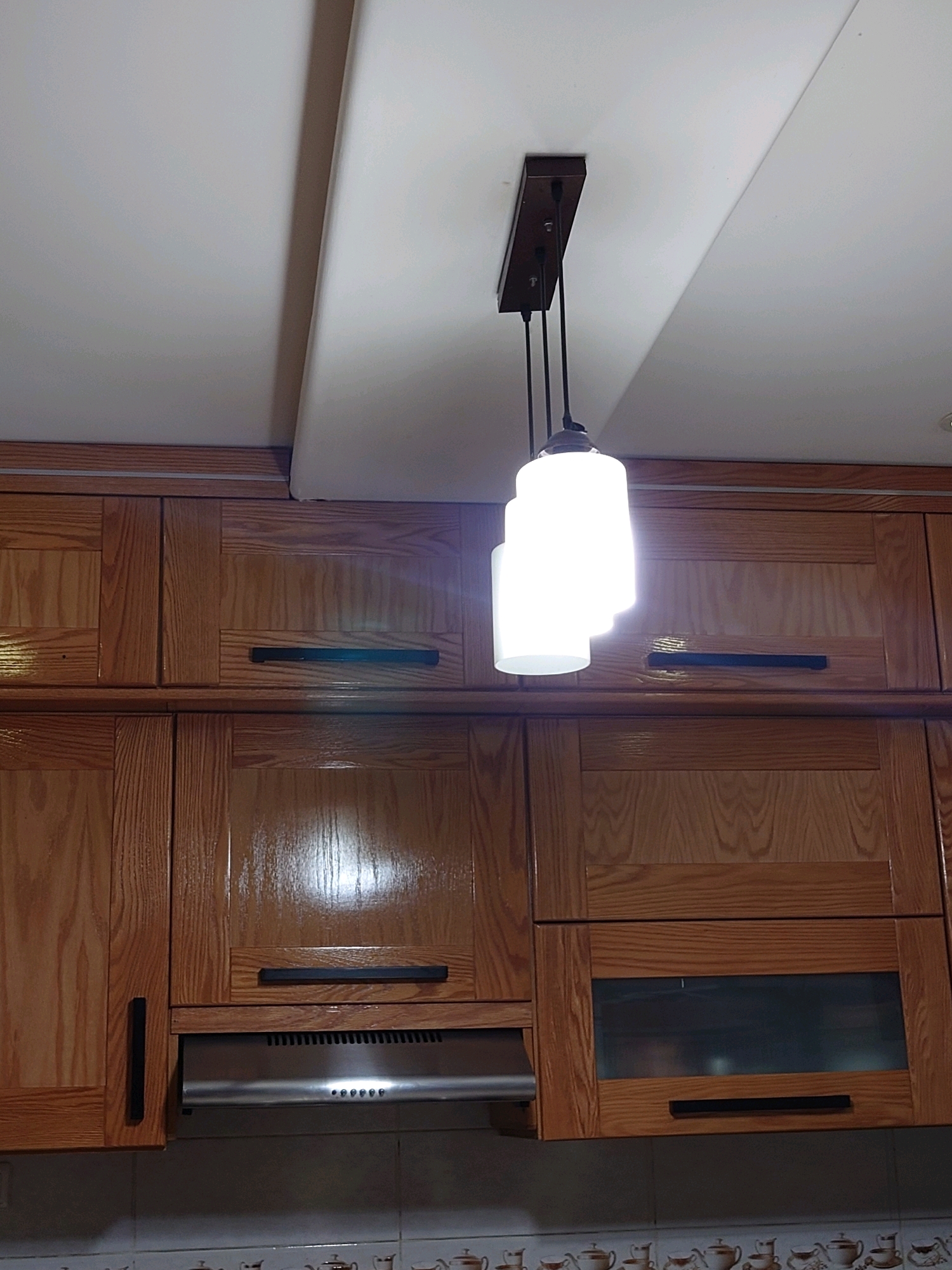 The image shows a close-up view of a kitchen. The cabinets are made of wood with a glossy finish and feature black horizontal handles. A stainless steel range hood is visible below one set of cabinets. Above the cabinets, a pendant light fixture with three white cylindrical shades hangs from a rectangular wooden base attached to the ceiling. The ceiling appears to be a flat white surface. The overall impression is of a functional, modern kitchen, likely in a residential setting within Ali Mendjeli, Algeria.