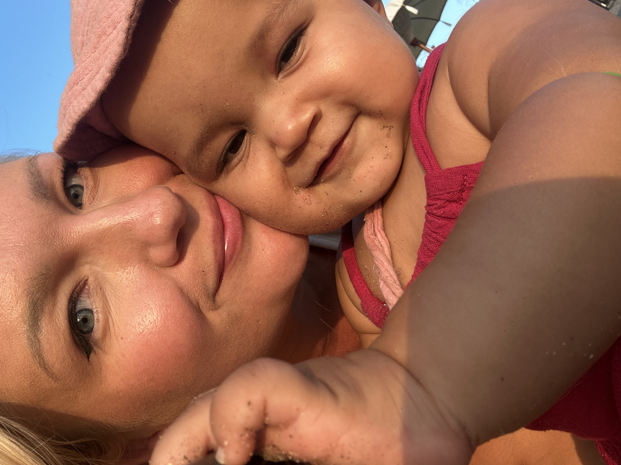 A heartwarming close-up captures an intimate moment between an adult woman and a baby, likely a mother and child, in North Kuta, Indonesia. The woman, with fair skin, blonde hair, and striking blue eyes, is shown in profile, leaning her head affectionately against the baby. Her expression is gentle and loving. The baby, with darker skin and dark eyes, wears a light pink bucket hat and a pink top, looking directly at the viewer with a wide, joyful smile.
Both individuals have visible grains of sand on their faces and arms, indicating they are at a beach or sandy outdoor location. The bright, natural light suggests a sunny daytime, with a warm glow on their skin. A clear blue sky dominates the background, with a subtle hint of a beach umbrella or canopy in the upper right corner. The image conveys a strong sense of happiness, affection, and bonding, capturing a tender moment of family enjoyment in a sunny, tropical setting. No text is visible in the image.
