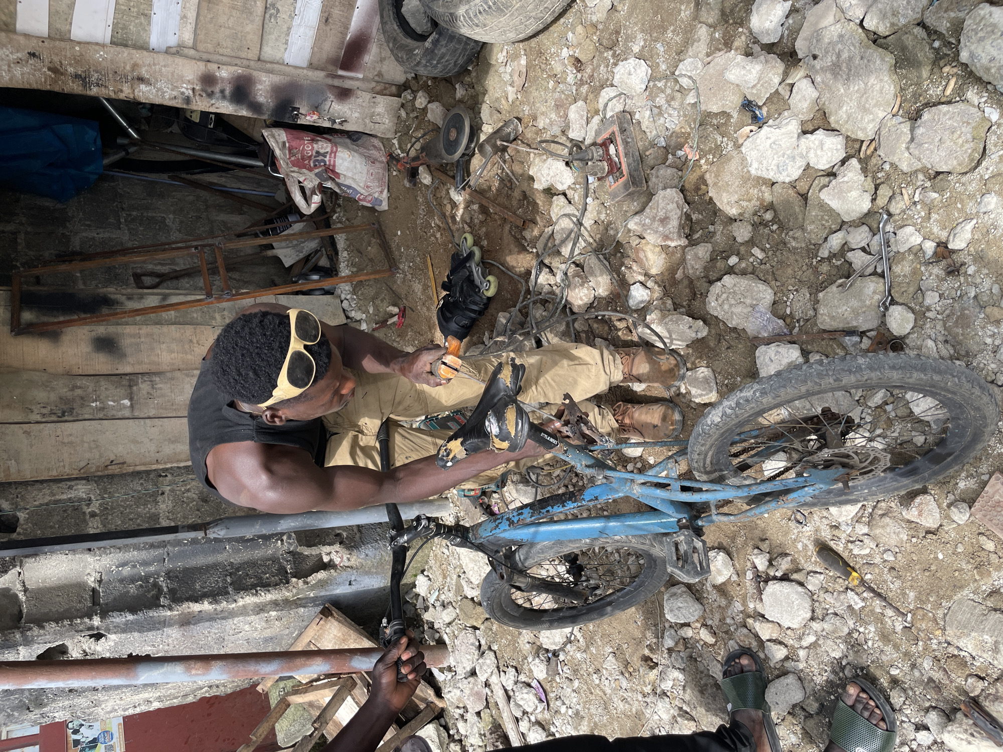 A Black man wearing a black tank top, khaki pants, and yellow-rimmed sunglasses is working on a blue bicycle in a cluttered outdoor environment. He is holding a tool near the bicycle's handlebars and appears to be making adjustments. The ground is covered with rocks and dirt. Various objects are scattered around, including rollerblades, an angle grinder, a tire, and a bag of cement. In the background, there is a dilapidated wooden structure with some cinder block walls. The overall scene suggests a workshop or repair area in an informal settlement. The image is captured from a slightly elevated angle, looking down at the man and the bicycle.