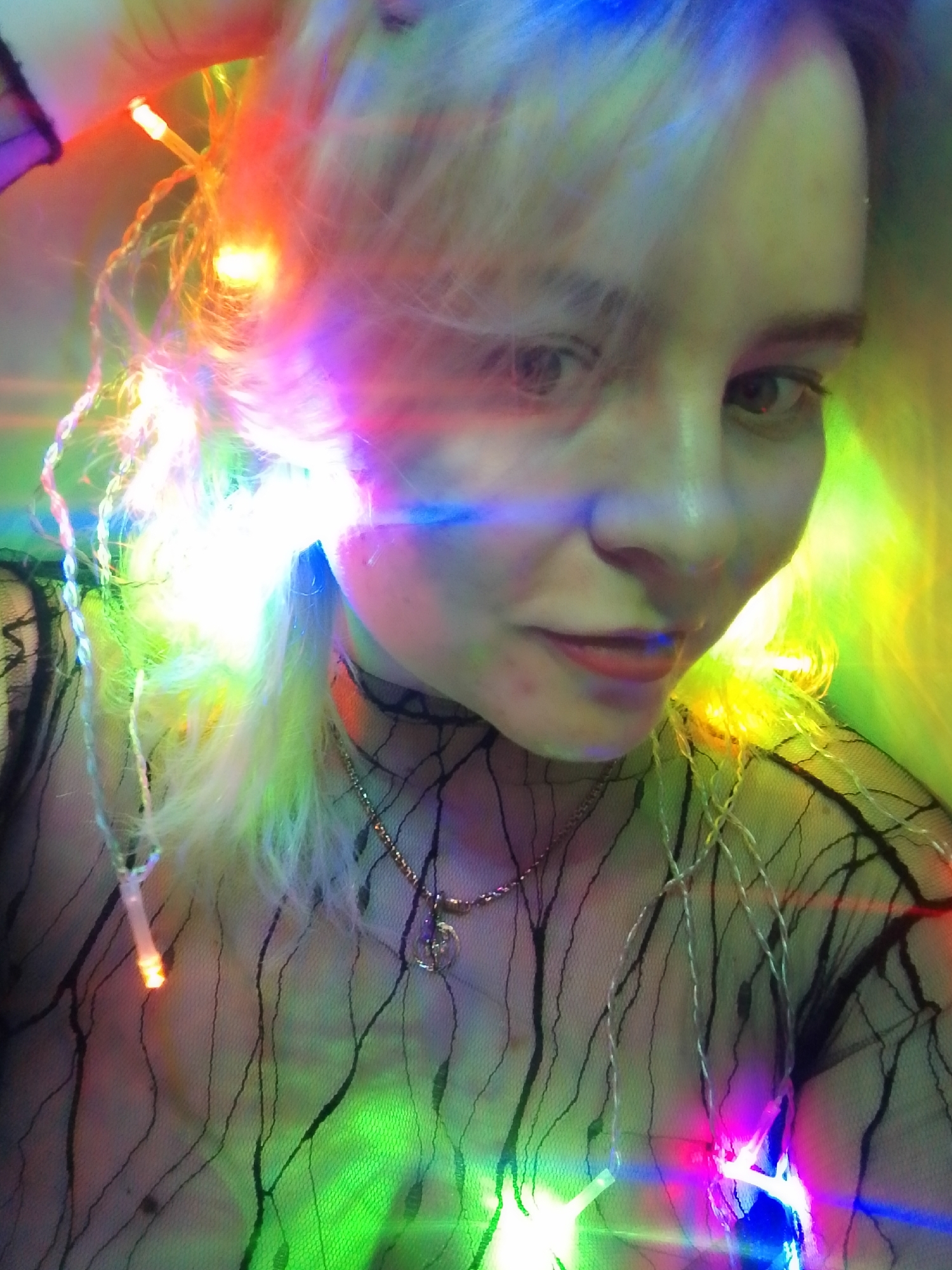 A close-up selfie of a person with light blond hair, lit by multicolored fairy lights. The person has fair skin, and is wearing a black, sheer, patterned top, a black choker, and a gold necklace. The fairy lights are interwoven in their hair and draped around their body. The lighting is intense and colorful. The location, Izhevsk, Russia, cannot be determined from the image.