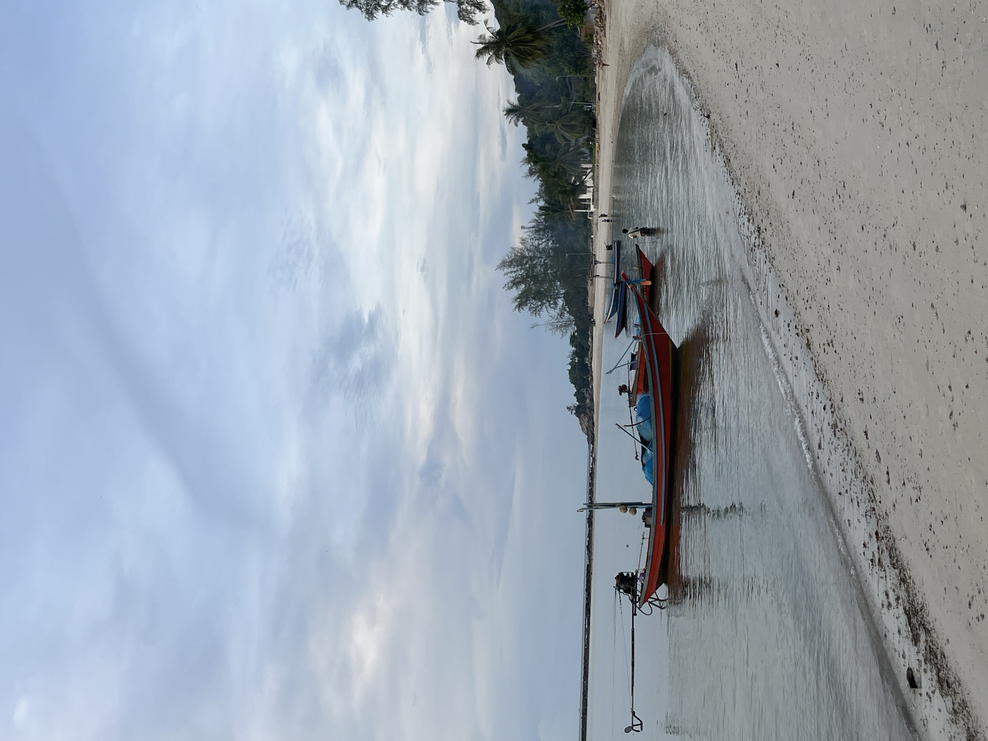 This image captures a tranquil beach scene in ตำบลเกาะพะงัน, Thailand, under an overcast sky. The upper two-thirds of the frame are dominated by a soft, light blue-grey sky with diffuse clouds, indicating an overcast day or an early morning/late afternoon. The horizon line is visible in the mid-distance.
The foreground and midground are characterized by calm, shallow water on the left, which gently laps against a wide, light-colored sandy beach that curves from the lower-right into the midground. The water's surface is smooth, reflecting the muted tones of the sky, with a thin line of white foam marking the ebb and flow of small waves at the shoreline. The sandy beach is a light beige-grey, textured with scattered small pebbles and debris, particularly closer to the water's edge.
Three traditional long-tail boats are positioned in the shallow water on the left side of the frame. The most prominent boat in the foreground is red with blue trim, featuring a long stern pole extending backward, characteristic of its propulsion system. Buoys are attached to a vertical pole near its bow. Partially visible behind it, further left, is another red boat. A third, darker blue or black boat is situated further out in the midground.
The coastline, forming the background of the beach on the right, is densely covered with tropical vegetation, including numerous palm trees and other broadleaf trees. Scattered among the trees in the midground-right, several white buildings with dark roofs are partially visible. In the far-left background, a long, dark, rocky breakwater or reef extends horizontally across the water.
At least four individuals are present in the scene. One person is standing waist-deep in the shallow water in the midground, to the right of the main boats, facing away from the camera. Two smaller figures are visible further down the beach on the right, one walking on the sand and another closer to the water's edge. A fourth faint figure can be discerned near the treeline in the background on the right. The lighting is soft and even, with minimal harsh shadows, consistent with an overcast environment.