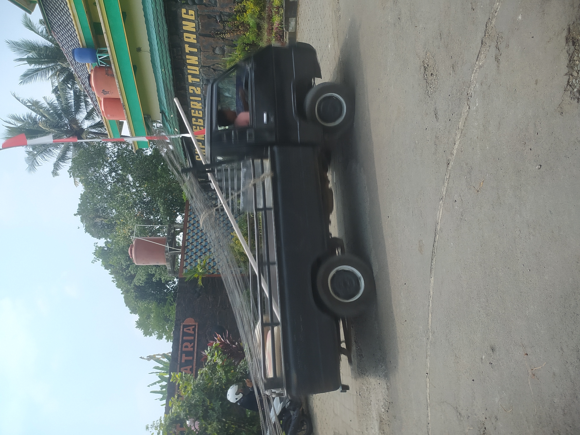A black pickup truck is parked on a paved road in front of a building with a green trim and a stone facade. The truck is carrying a load of metal rods and a white pipe in its bed. A person is visible in the driver's seat of the truck. Behind the truck, a sign on the building reads "SMP NEGERI 2 TUNTANG". The sky is clear and blue, with some trees and palm trees visible in the background. A red and white flag is flying from a pole in front of the building. The scene appears to be in the city of Candirejo, Indonesia, based on the building's signage.
