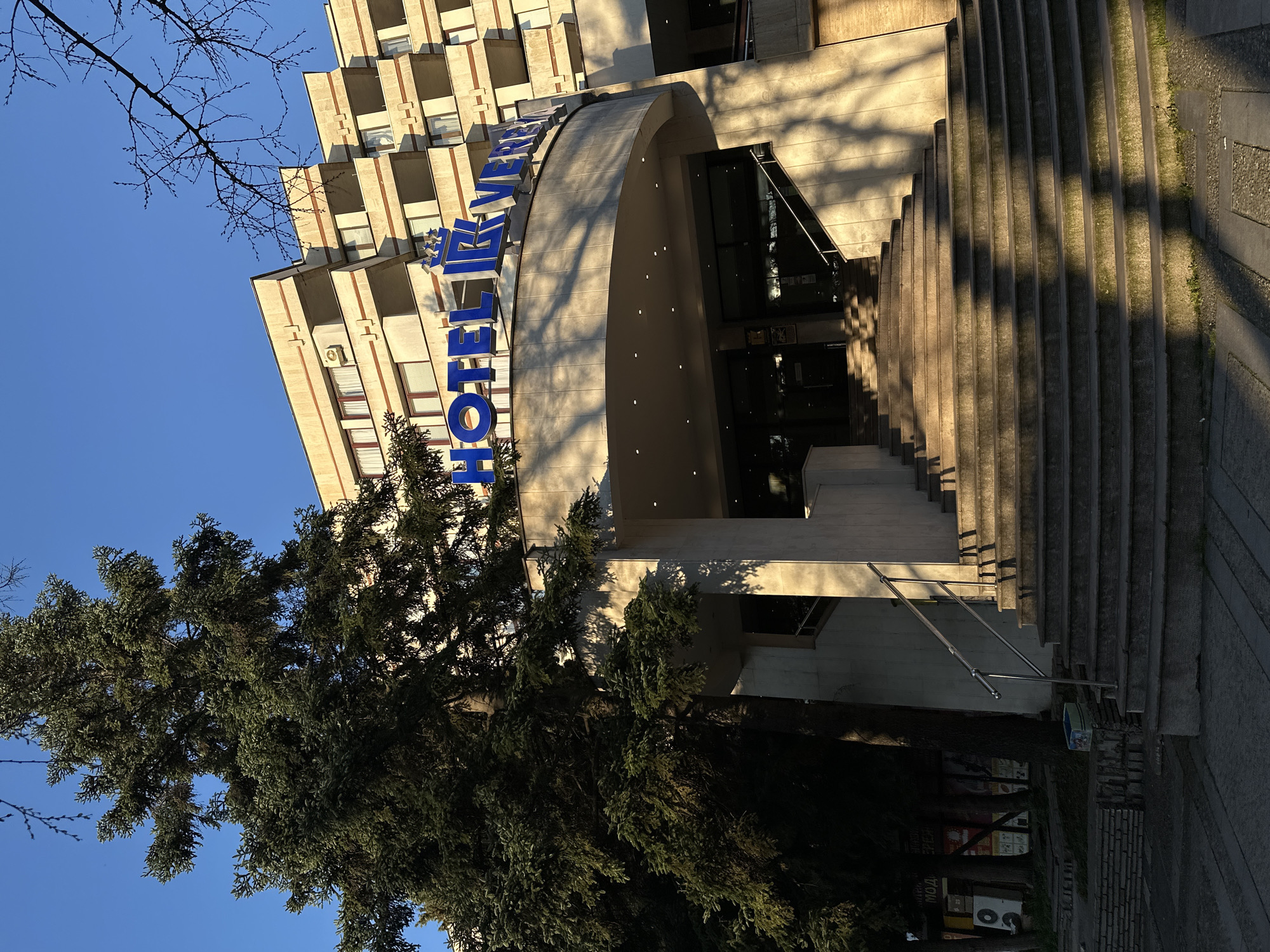 The image depicts the entrance of Hotel Rila in Stara Zagora, Bulgaria. The building is multi-storied with a concrete facade. The hotel's name is displayed in large blue letters above the entrance. A curved concrete awning covers the entrance, which is accessible via a wide set of concrete stairs. Shadows from nearby trees are cast across the building and stairs. A large evergreen tree is present in the foreground, partially obscuring the view of the hotel. The sky is clear and blue. A small part of the street is visible on the right-hand side.