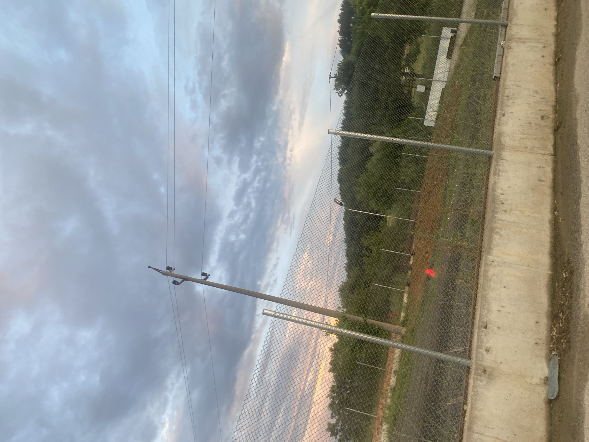 An overcast sky with dramatic clouds casts a muted light over a landscape dominated by a chain-link fence and a concrete barrier in the foreground. A wooden utility pole, supporting power lines, stands tall against the sky. Behind the fence, a wooded hillside rises, dotted with a few scattered buildings. The ground beyond the fence appears to be a mix of dirt and gravel, with some areas of sparse vegetation. Two small red markers are visible on the ground, and a larger white structure is partially obscured by trees. The scene suggests a transitional or undeveloped area, possibly on the outskirts of Ablanitsa, Bulgaria, given the overall visual characteristics.