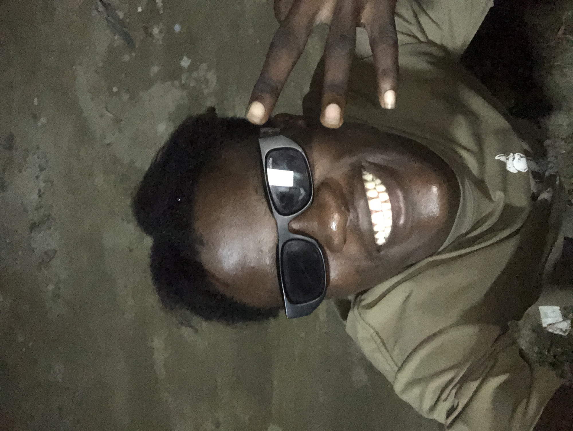 A medium shot captured from a slightly low angle shows a Black male, likely in his late teens or early twenties, posing for a selfie. He has dark, curly hair styled upwards and is wearing dark-rimmed sunglasses. A wide, toothy smile is visible, and his right hand is raised, displaying a peace sign. He is wearing a plain, olive-green t-shirt. The background is a textured, uneven surface, appearing to be a wall or ground with patches of lighter and darker tones, possibly indicating dampness or age. The lighting is dim, casting subtle shadows and highlighting the subject's features. Based on the context provided, this image was taken in the city of Ado, Nigeria.