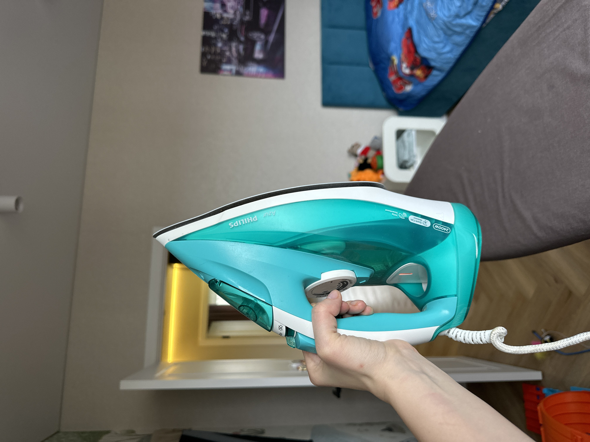 The image, taken in Chaikovsky, Russia, features a person's right hand holding a teal and white Philips Azur steam iron. The iron is positioned horizontally, with the hand gripping its ergonomic handle.
Visible text on the iron includes "PHILIPS," "Azur," "2400W," "Quick Calc Release," "OptimalTEMP," and "SteamBoost." The circular temperature dial is partially visible, indicating settings for various fabrics such as "LINEN," "COTTON," "WOOL," "SILK," and "SYNTHETICS." A white power cord with a coiled protector extends from the rear of the iron.
The background depicts an indoor residential setting, likely a bedroom or living space. A light-colored wall (beige or light gray) dominates, with a dark, blurred picture or poster hanging on it towards the left. To the right, a bed is visible, covered with a blue duvet featuring red patterns, possibly characters like Spider-Man. Next to the bed, a small white nightstand holds indistinct objects, possibly toys. A dark brown or gray blanket or mattress occupies the right side of the frame, suggesting another bed or sofa.
Below the iron, a white shelf or windowsill is visible, featuring a prominent yellow-orange glow from what appears to be an illuminated window frame, suggesting daylight. Part of an orange container is also discernible on a lower surface in the bottom right corner. The scene captures a domestic moment, with the iron being held but not actively in use for ironing. The overall image has a tilted orientation.