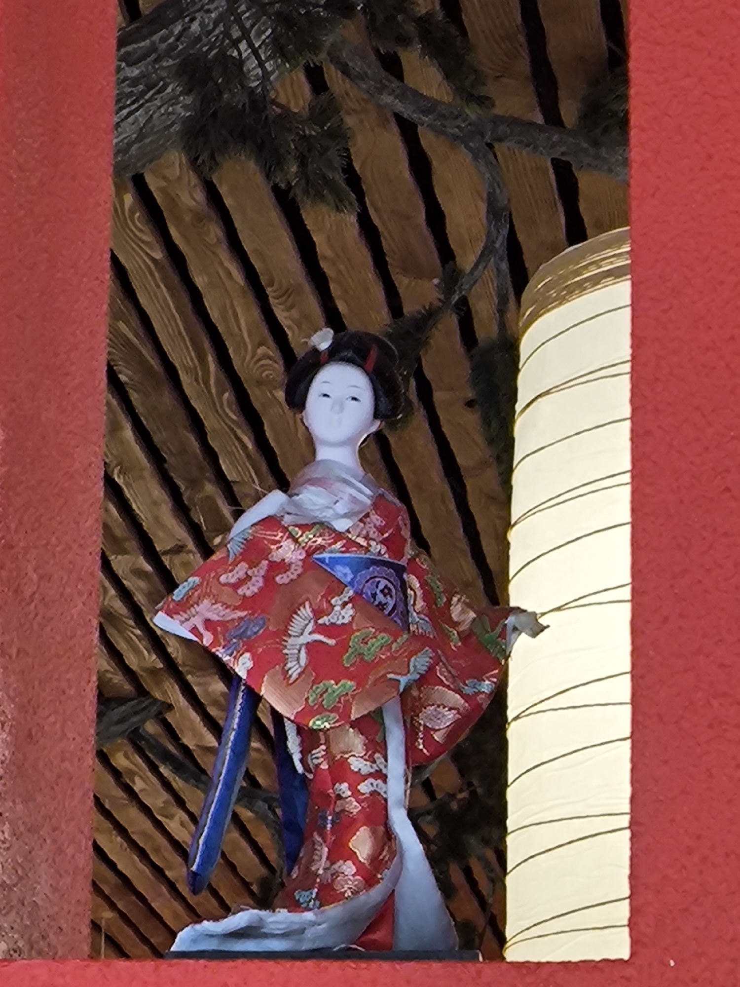 A geisha doll is on display in a decorative niche. The doll wears a red kimono with a crane pattern. The niche has a wood slat background and a red border. A cylindrical paper lantern is to the doll's right. The image was taken in the city of Moscow, Russia.
