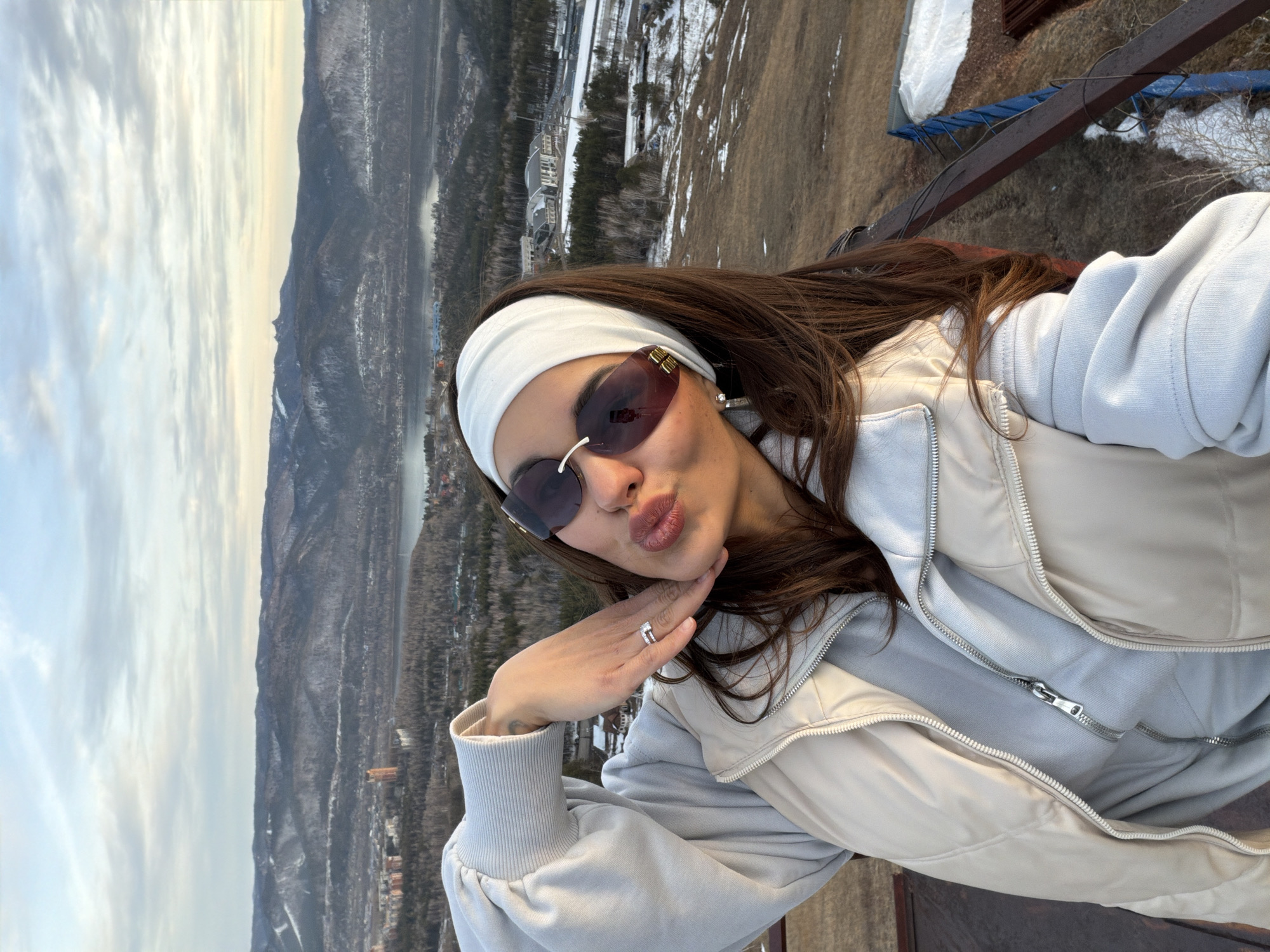 A woman in sunglasses and a headband takes a selfie, puckering her lips slightly and resting her chin on her hand. She wears a light-colored jacket and sweatshirt. The background shows a panoramic view of Krasnoyarsk, Russia, with mountains, a river, buildings, and forest visible under a cloudy sky. The woman is on a structure made of metal.