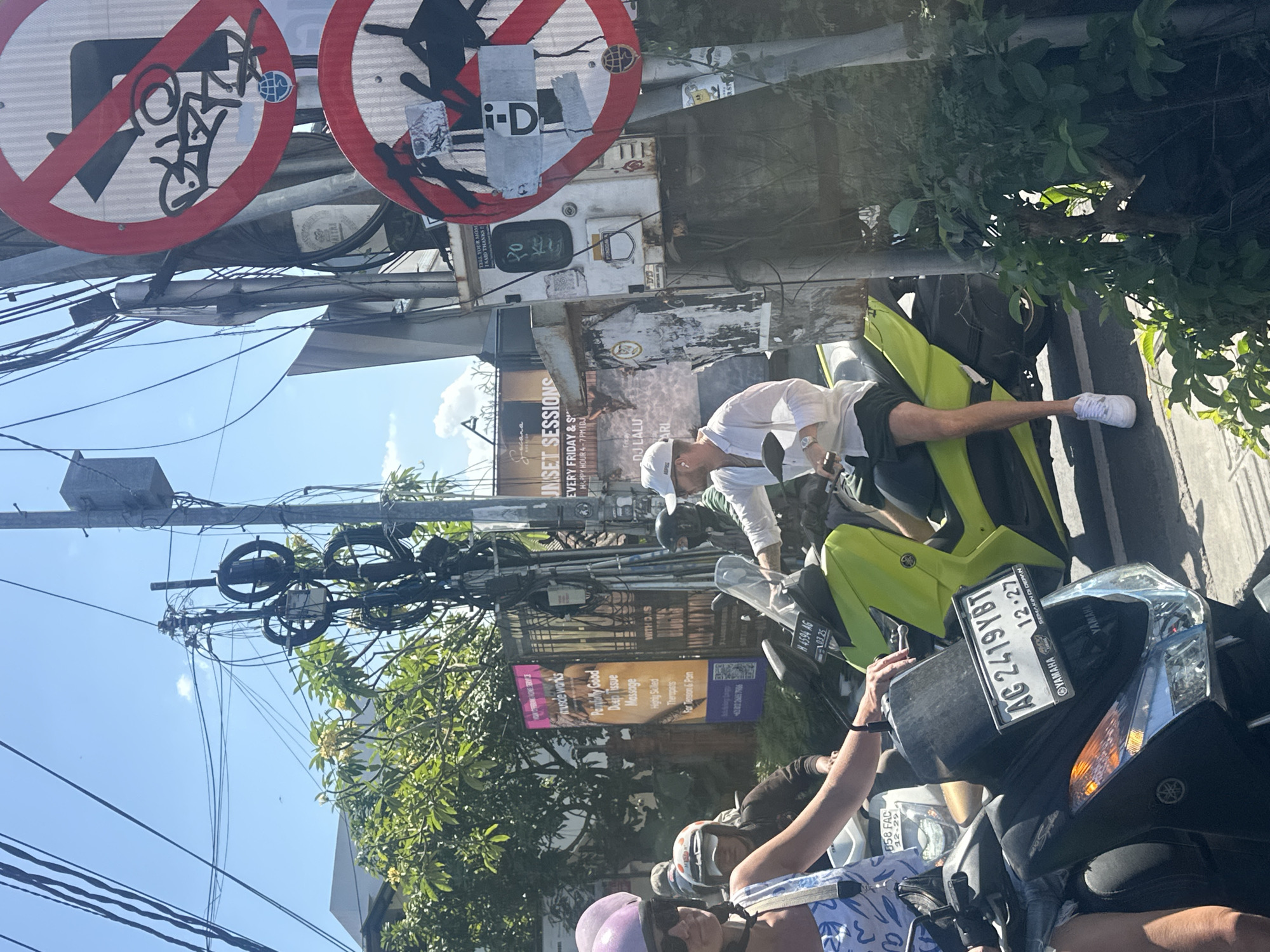 The image captures a street scene in North Kuta, Indonesia, on a sunny day, indicated by the strong shadows and bright light. A variety of motorcycles are parked and in motion. In the foreground, a person wearing a white long-sleeved shirt and dark shorts is seated on a bright lime green scooter. They are wearing a white baseball cap and white sneakers. To the left, a woman in a blue and white patterned top and a light purple helmet is partially visible, also on a scooter. Several other individuals on motorcycles are present in the background, some wearing helmets.
Traffic signs, one with a red circle and a diagonal line across a left turn symbol, and another with a similar prohibition symbol over what appears to be a motorcycle, are visible on a pole adorned with numerous electrical wires and cables, suggesting a busy urban environment. Posters advertising services, including "muscle works Painfully Good Deep Tissue Massage" and "Sunset Sessions," are plastered on a wall behind the scooters. The license plate of the lime green scooter reads "AG 2419 YBT" with a date "12-27". The overall impression is one of everyday life on the streets of North Kuta, with traffic, advertisements, and infrastructure intermingling.