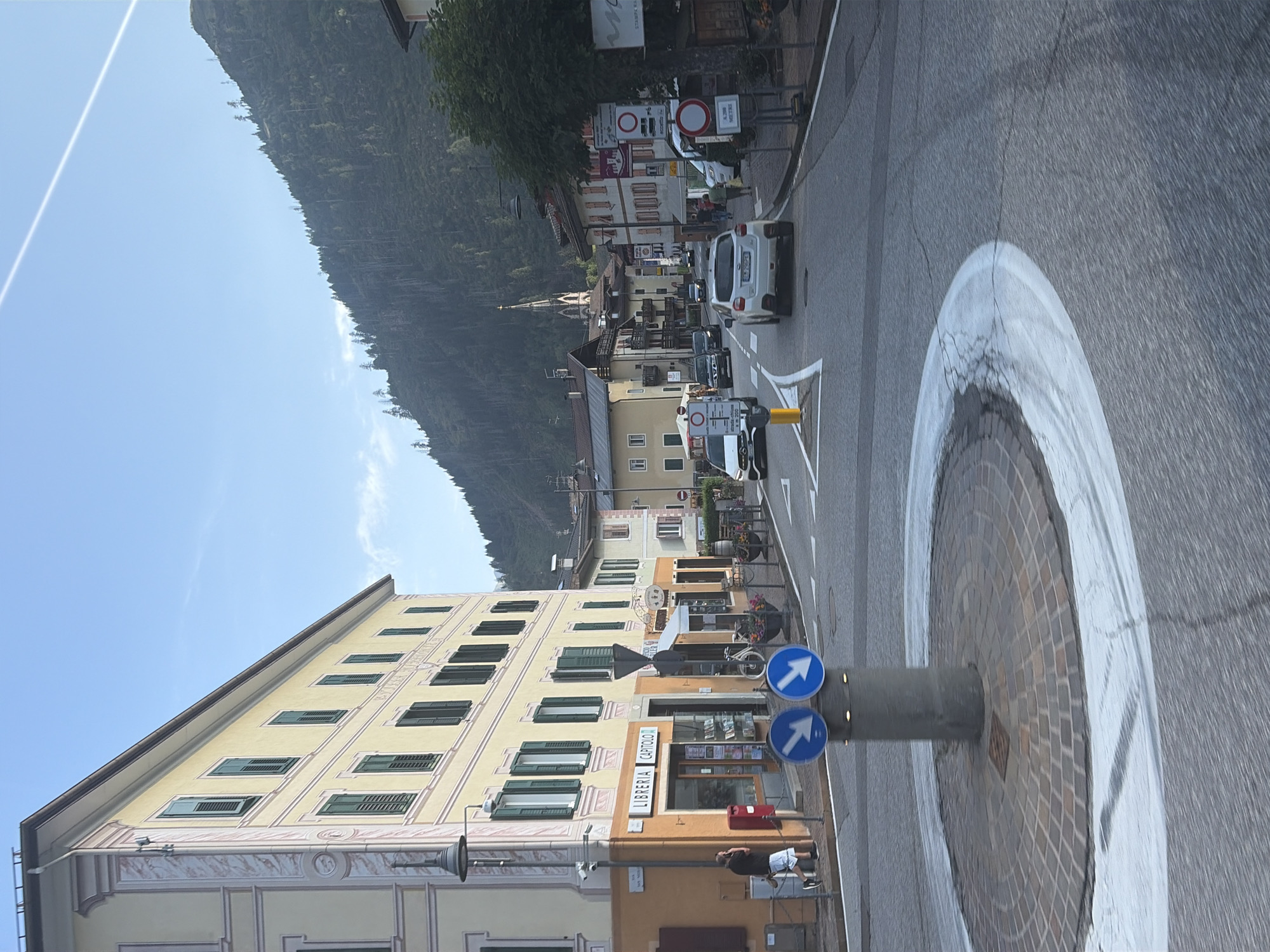 The image captures a street scene in Predazzo, Italy, likely on a clear day with a bright blue sky and a faint contrail. In the foreground, a circular roundabout is visible, paved with stone tiles and outlined by white markings on the asphalt road. A gray cylindrical pole stands in the center of the roundabout, topped with two circular blue signs, each displaying a white downward-pointing arrow.
To the left of the roundabout, a multi-story building with a pale yellow facade and decorative pink trim dominates the view. The building features numerous windows with dark green shutters. At the base of this building, a shop with a sign reading "LIBRERIA CAPITOLO" is visible, with books displayed in its window. A red mailbox is positioned next to the shop entrance. A man in a dark shirt and shorts stands near the building, appearing to be on his phone.
The street itself is paved with asphalt and shows signs of wear, with white lane markings and some visible tire marks. Several cars are present on the road, including a white SUV in the middle lane and other vehicles further down the street. Buildings line the opposite side of the street, featuring a mix of architectural styles and colors, with some exhibiting pitched roofs. A church spire with a cross is visible in the distance, rising above the rooftops.
The background of the image is dominated by a steep, tree-covered mountainside. The trees are dark green, creating a dense forest that stretches up the slope. The overall scene depicts a typical European town street, with a blend of residential and commercial buildings set against a natural mountain backdrop.