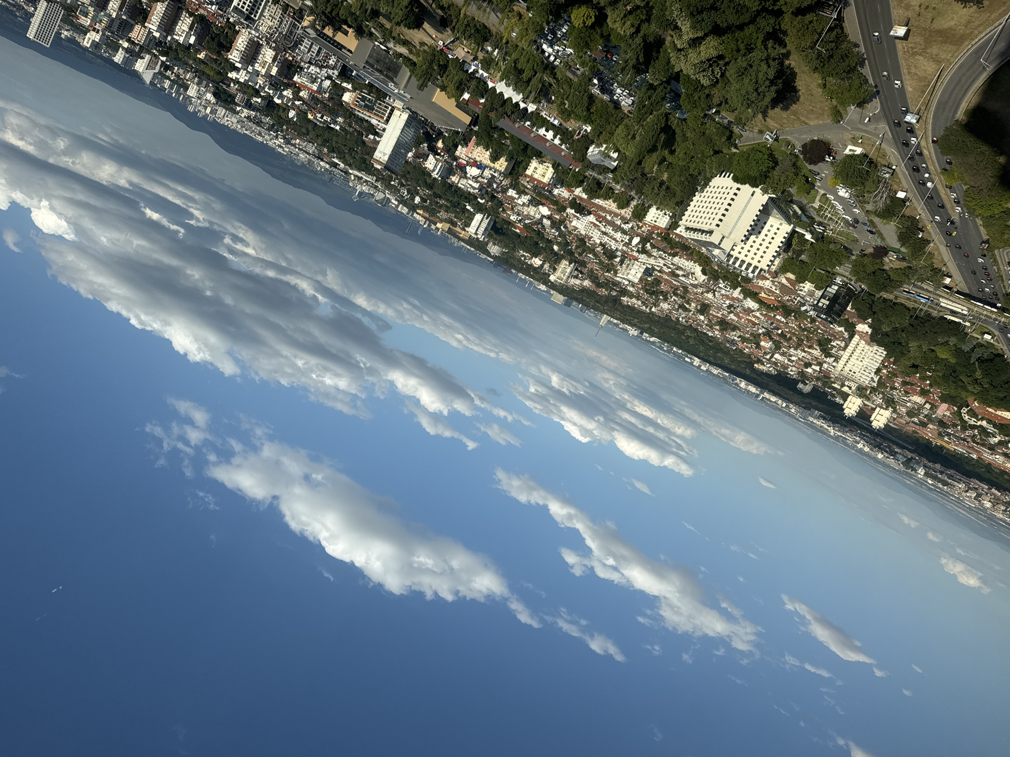 The image captures an aerial view of Sofia, Bulgaria, tilted at an angle. The sky is a vibrant blue, dotted with scattered white and gray clouds. Below, the city unfolds with a dense arrangement of buildings, a mix of residential and commercial structures, many with red-tiled roofs. Lush green trees are interspersed throughout the urban landscape, forming pockets of foliage. A highway with moderate traffic, including cars and buses, curves through the lower portion of the scene. In the distance, a range of mountains forms a hazy backdrop against the horizon. The scene depicts a typical day in a large European city, with a blend of architecture, nature, and infrastructure under a partly cloudy sky.