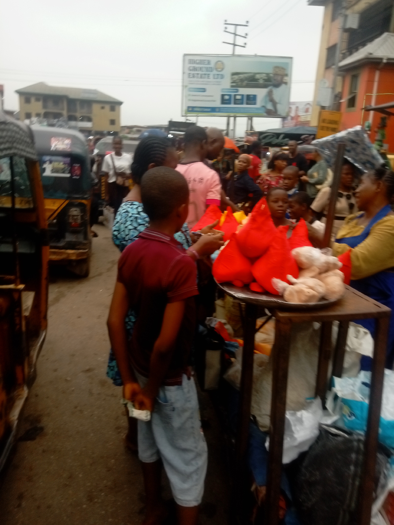 On a cloudy day in Onitsha, Nigeria, a bustling street scene unfolds. A young boy in a red shirt and light-wash shorts stands near a makeshift food stall. The stall, constructed from dark metal, displays red cone-shaped items alongside pale, likely skinned, chicken. An adult woman in an apron stands behind the stall, possibly selling the items. A crowd of people surrounds the stall, some interacting with the vendor, others simply passing by. Vehicles, including a yellow-orange "Keke" (auto rickshaw), navigate the crowded street. In the background, buildings of varying heights are visible, along with a billboard advertising real estate.