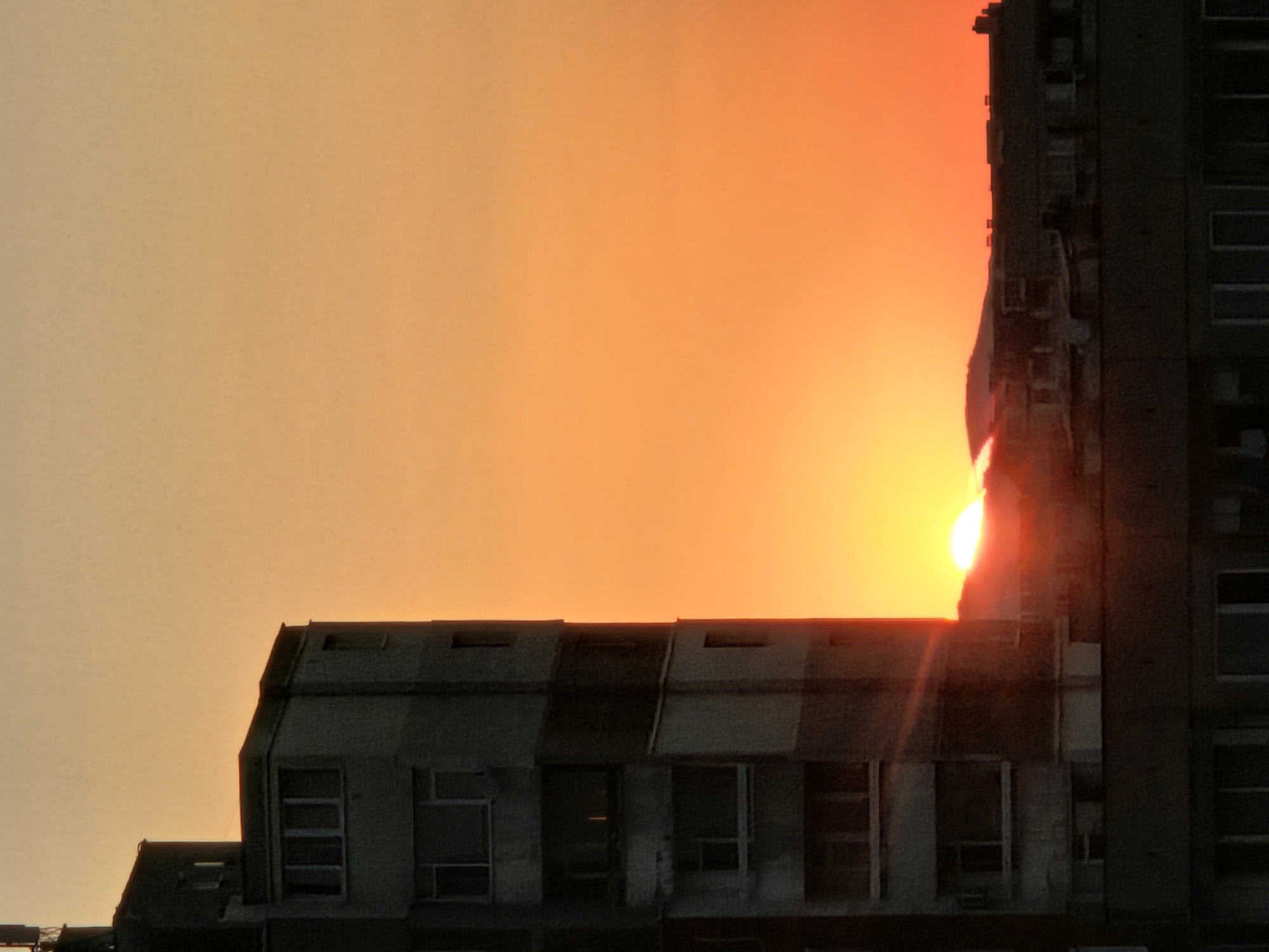 The image depicts a sunset over a city skyline. A multi-story building, characteristic of Soviet-era architecture, stands prominently on the left side of the frame. The building is grey and appears to have balconies and numerous windows. In the background, the sun is setting behind a silhouette of buildings and a hill or mountain range. The sky is a vibrant gradient of orange and yellow, indicating the time of day is either sunrise or sunset. The scene is devoid of any visible people or specific activities. The setting appears to be an urban environment, consistent with a location in Sofia, Bulgaria, given the architectural style of the buildings.