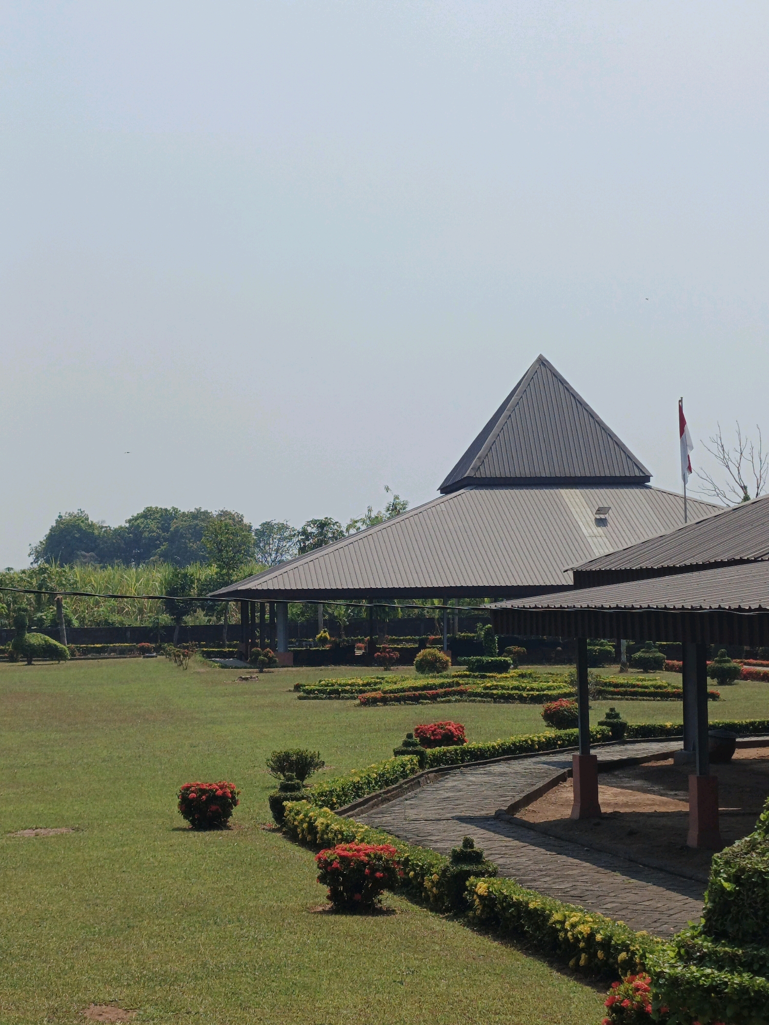 The image depicts an outdoor scene at a park or garden in Kejagan, Indonesia, under a clear, pale blue sky. The foreground features a well-maintained lawn with several small, rounded bushes of red flowers. A paved walkway, constructed from interlocking rectangular stones, winds through the scene, bordered by manicured hedges and more flowering bushes, some with red and yellow blossoms.
In the mid-ground, the park contains a series of covered gazebos with dark, sloped roofs. One larger structure in the background has a distinctive pyramidal roof, topped with a metal ridge. A flagpole with the Indonesian flag (red and white) is visible near this larger structure. The landscaping includes rows of neatly trimmed bushes and small trees.
Beyond the manicured garden, a dense line of trees and tall green plants, possibly sugarcane or bamboo, forms a backdrop. The overall environment appears peaceful and well-kept, suggesting a public recreational space. No people are visible in the frame, and no specific actions or interactions are taking place. The lighting suggests it is daytime, likely morning or late afternoon, given the soft shadows.