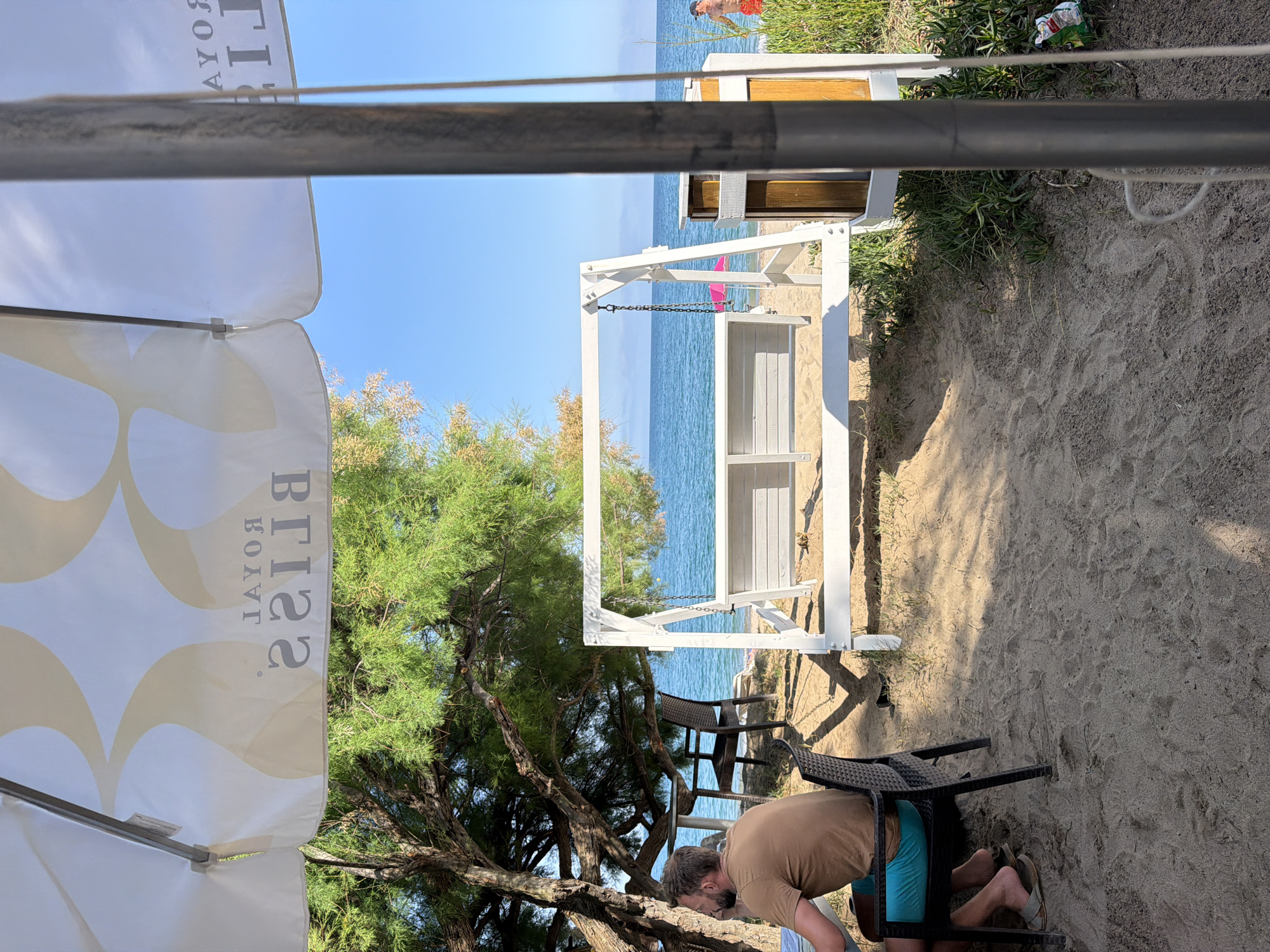 The scene depicts a beach setting near Mont-roig del Camp, Spain. The view is partially obstructed by a white sun umbrella with the reversed text "ROYAL BRISA". The beach is sandy, with vegetation interspersed, leading towards a view of the blue sea. A white, wooden lifeguard tower with two visible levels is present. A man wearing a brown t-shirt and blue shorts is crouched near the base of a black patio table. Additional patio chairs are situated near the base of a tree, which provides shade. A discarded trash item is seen on the sand near the lifeguard tower.
