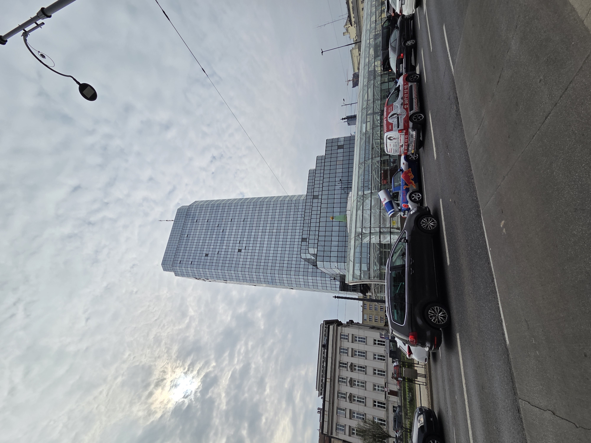 A street scene in Warsaw, Poland features a tall, modern glass skyscraper rising vertically. To the right, a line of parked cars partially obstructs the view of a lower, older building with a traditional facade. The sky overhead is cloudy, with sunlight partially visible. A street light is visible on the left.
