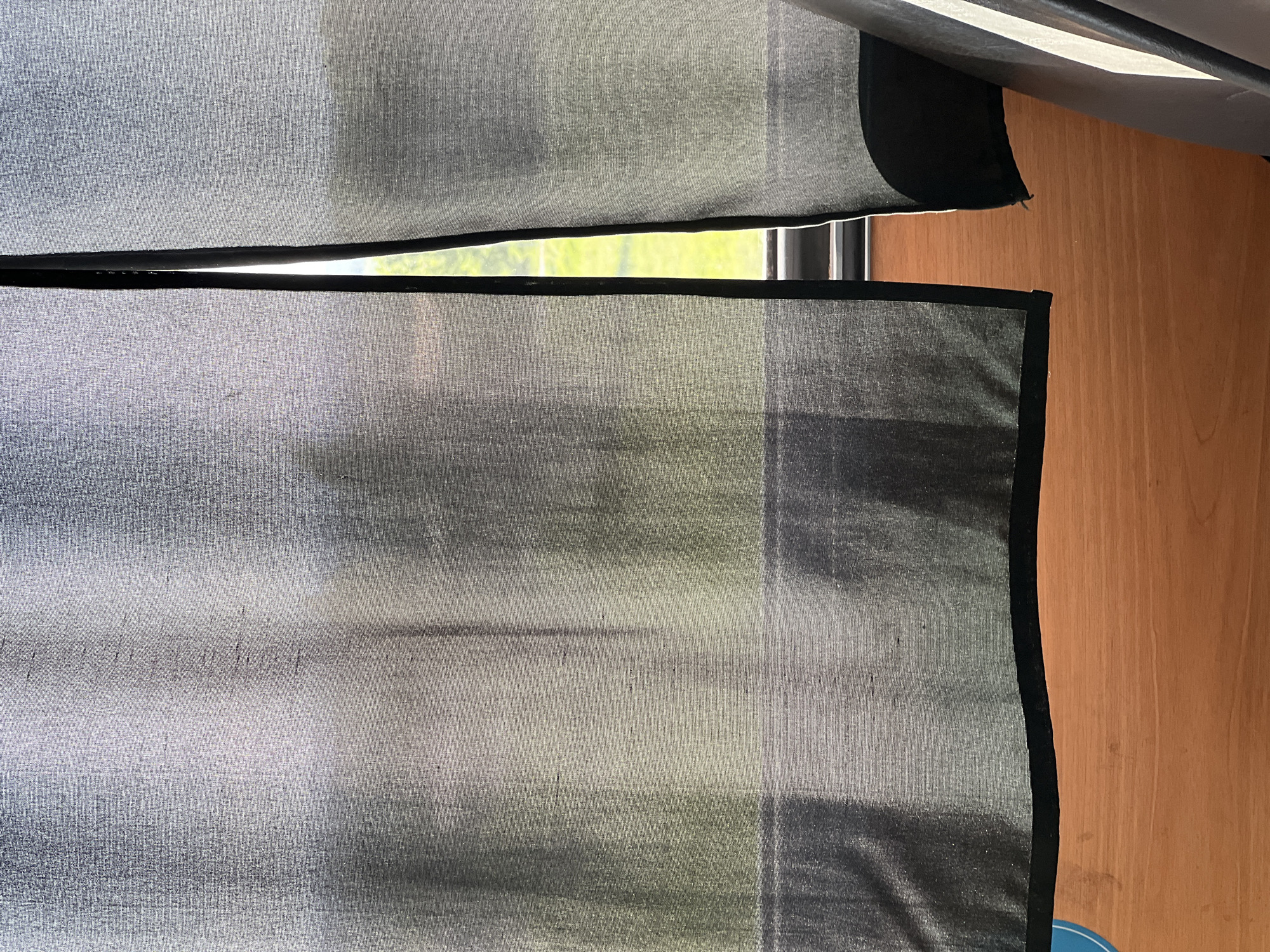 The image shows a close-up of a window covered by two sheer, gray curtains with black trim. A sliver of green foliage is visible through the partially open window. To the right of the curtains is a light brown, wood-paneled wall. The angle of the shot suggests the viewer is positioned slightly below the window. There are no people visible. The location in the city of Бизяки, Russia is not visually identifiable from the image.