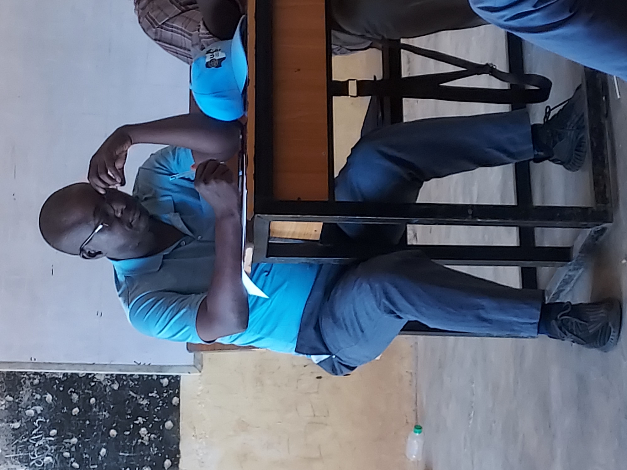 A man in a light blue shirt and dark trousers sits at a desk, his head resting on his hand. A blue cap is on the desk next to him. The setting appears to be a classroom or office space, and the location is the city of Jos, Nigeria.
