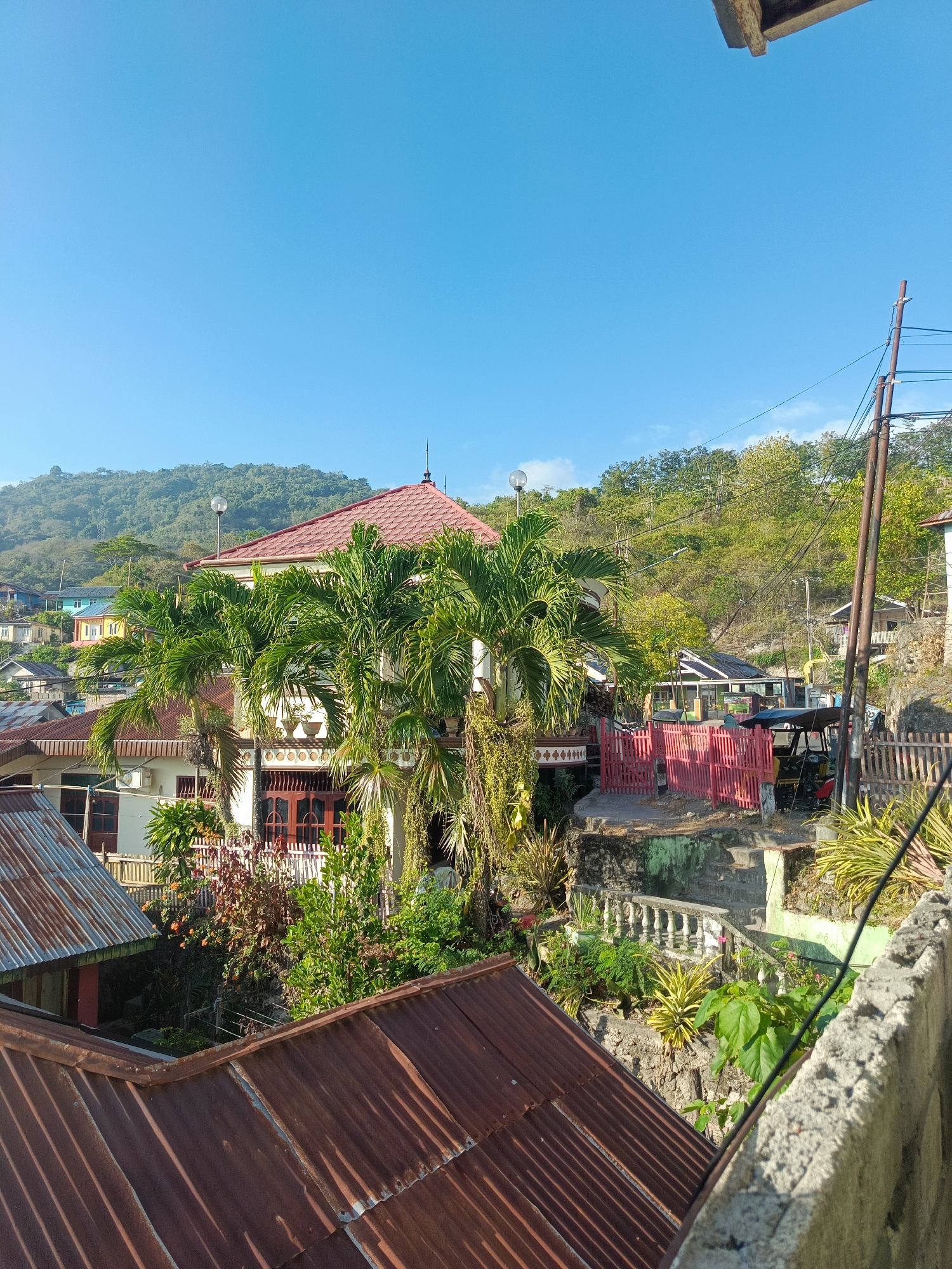 The image depicts a residential area with a clear blue sky overhead. The scene is characterized by a mix of traditional and modern housing, with corrugated metal roofs dominating the foreground. Lush green vegetation, including palm trees, is interspersed throughout the landscape, suggesting a tropical environment. In the mid-ground, a two-story house with a red tiled roof and white walls stands prominently, adorned with decorative elements. Adjacent to it, a bright pink fence encloses a courtyard where a small three-wheeled vehicle, possibly a tuk-tuk, is parked. The background features a gently sloping hillside covered with trees and smaller houses, indicating the hilly terrain of the region. The overall impression is that of a sun-drenched day in a densely populated, somewhat rustic neighborhood. The presence of utility poles and wires suggests a functional infrastructure within the community. While the image focuses on the built environment and natural elements, there is no indication of specific landmarks or activities that would pinpoint a precise location within the city of Gorontalo, Indonesia.