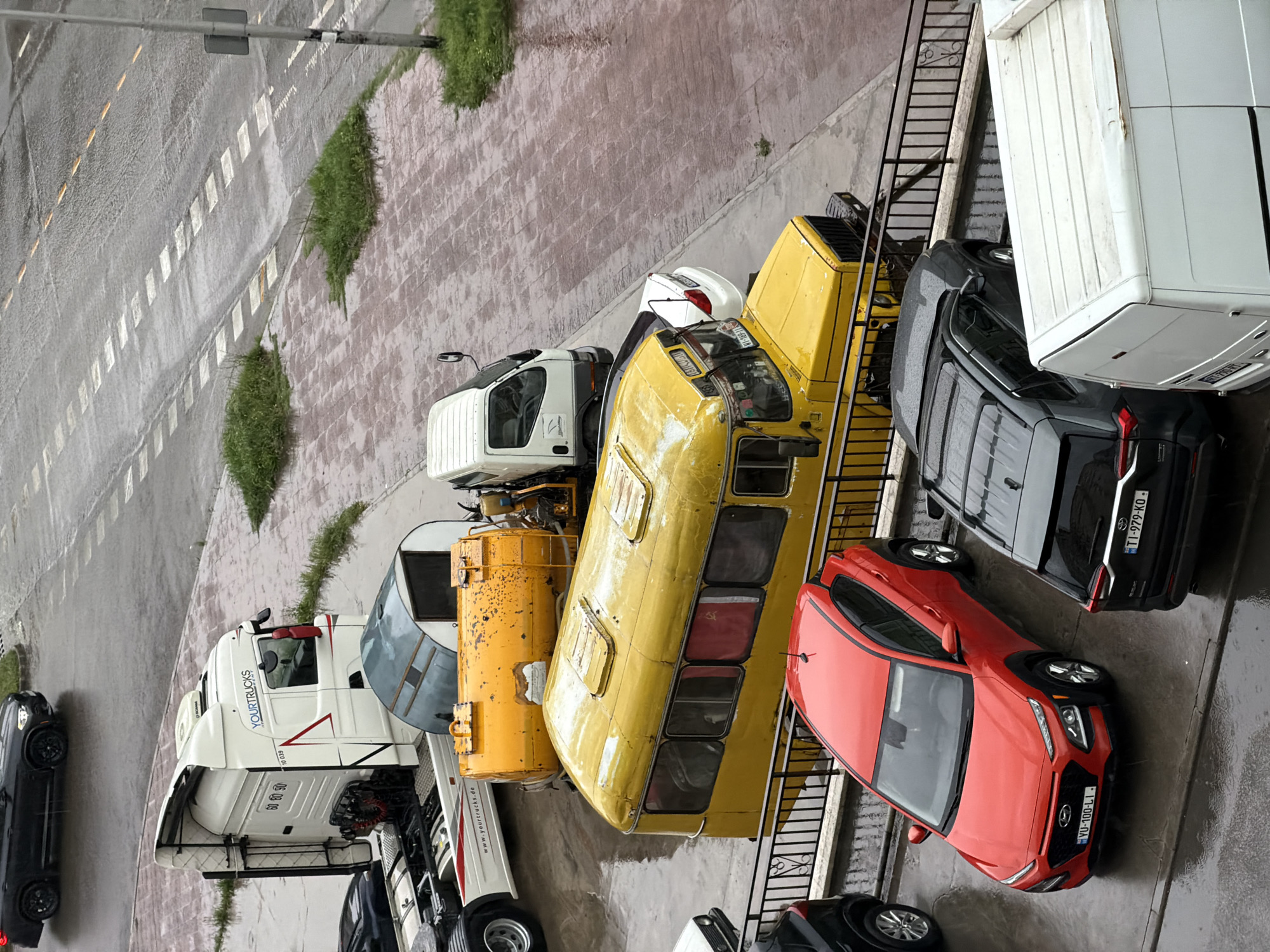 A high-angle shot shows a diverse array of vehicles parked on a wet surface, possibly in a commercial or industrial area. The setting appears to be in Tbilisi, Georgia, based on the license plates visible on the vehicles.
Prominent among the vehicles are a large white semi-truck with the brand "YOURTRUCKS" and a partially visible European license plate ending in "10 039," a yellow vehicle that resembles a repurposed bus or industrial vehicle, a red Hyundai Kona with Georgian license plate "YU-100-LI," and a black SUV with Georgian license plate "TI-979-KO." There are other trucks and vans, including one that appears to be a box truck, parked closely together.
The surface is wet, reflecting light and indicating recent rainfall. The area seems to be paved, with some sections showing what could be painted markings or pedestrian walkways. Vegetation is visible along the edges of the paved area.