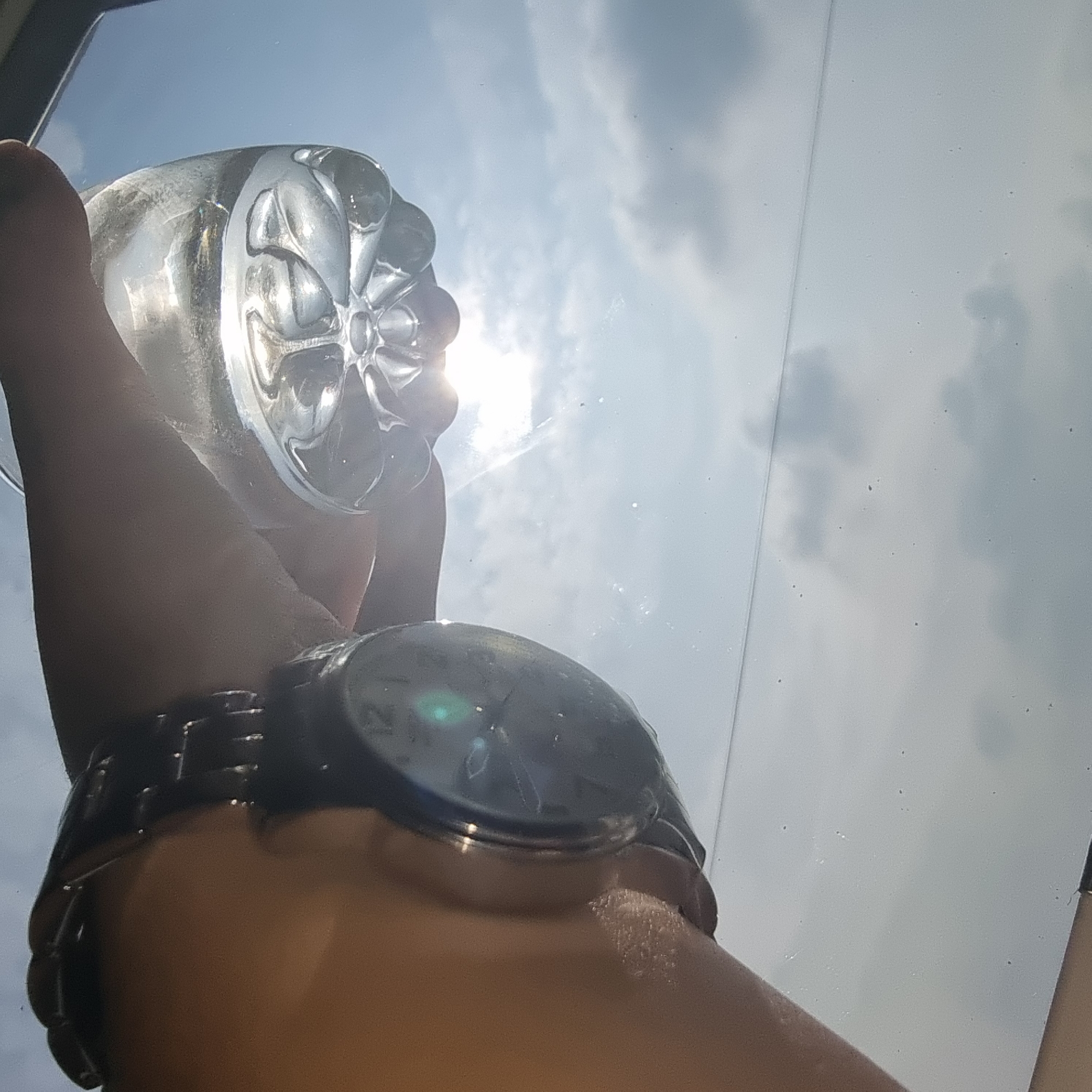 The image shows a close-up, low-angle view of a person's wrist wearing a dark watch, holding a clear plastic water bottle filled with water. The bottle is held up towards a bright sky, causing the water and bottle to refract the sunlight. The background consists of a bright, hazy sky with some scattered clouds. The scene appears to be outdoors, possibly in a location with ample sunlight. No specific details of the city of Dunnae-myeon, South Korea are discernible from this image. The image is a snapshot of an everyday object interacting with natural light, emphasizing the reflective and refractive properties of the water and plastic.