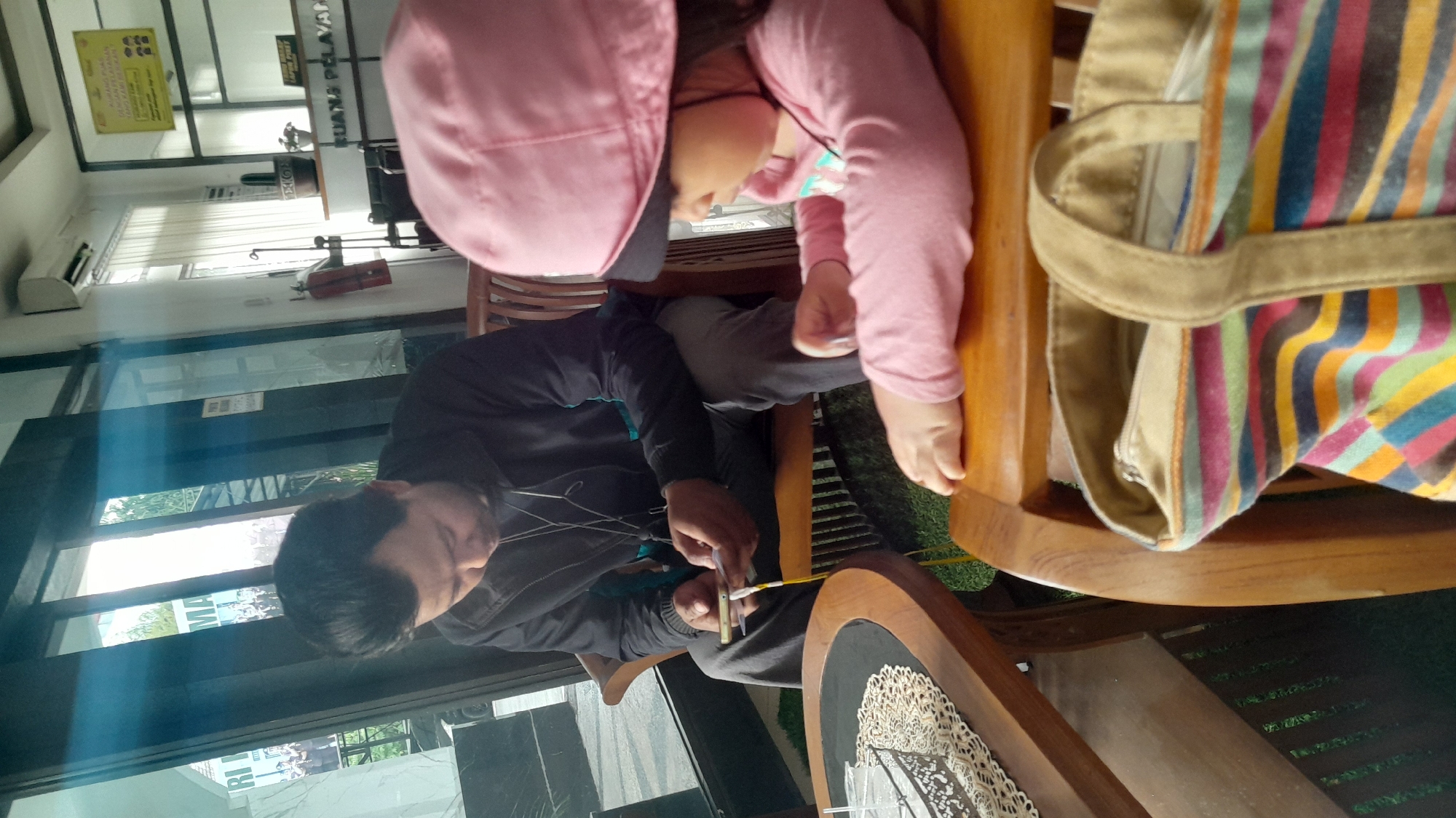 The image shows a man and a young child sitting at tables in what appears to be an indoor cafe or restaurant in Tasikmalaya, Indonesia. The man is wearing a dark jacket and grey pants and is looking down at his phone. The child is wearing a pink top and a pink hat, and is looking towards the man. A striped bag with a canvas strap is visible in the foreground, resting on a wooden chair. In the background, a fire extinguisher is mounted on the wall. The cafe has large glass windows, revealing an outdoor area with greenery. Signs are visible in the background, one of which reads "RUANG PELAYANAN" which translates to "SERVICE AREA".
